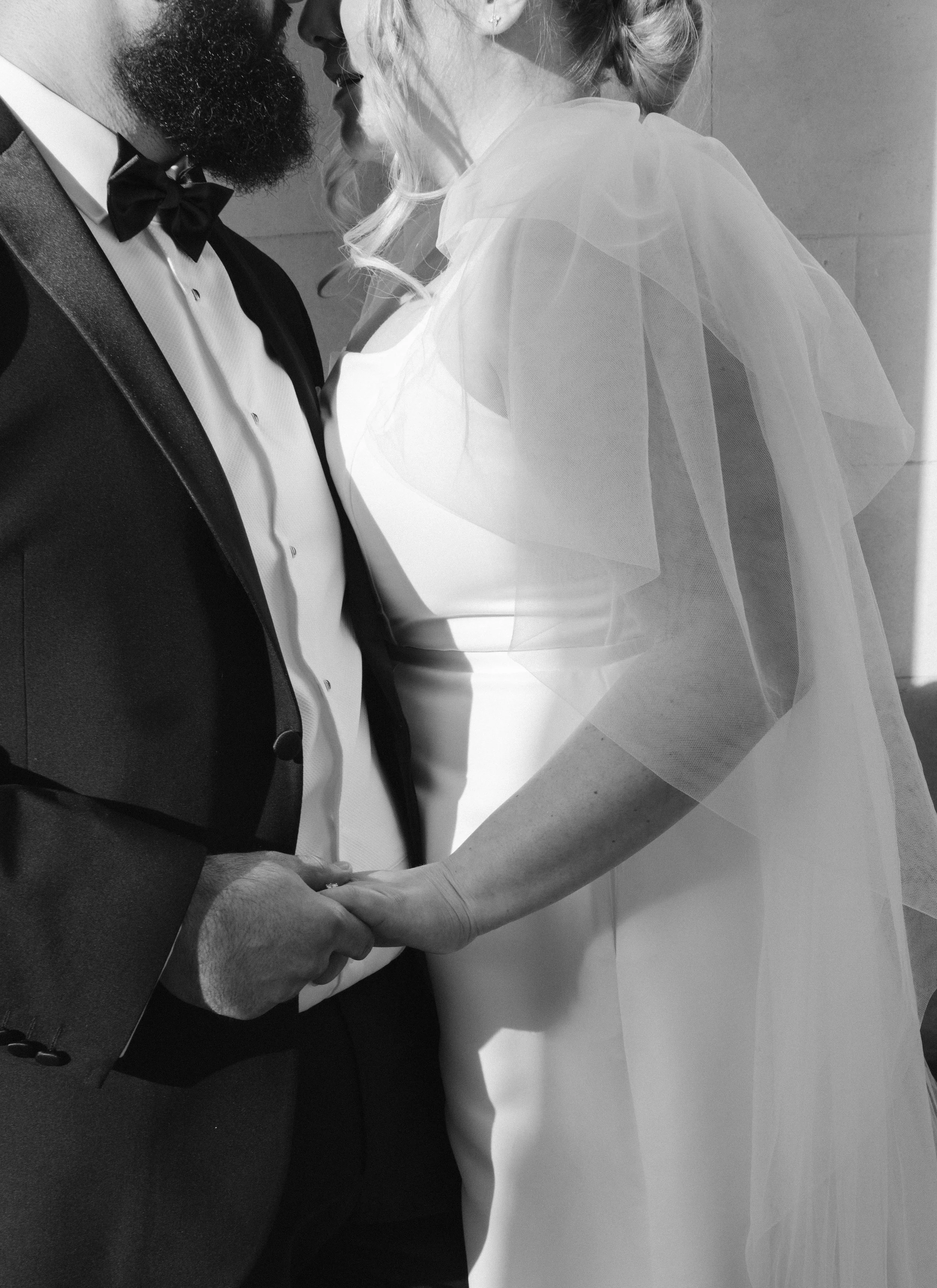 A close-up of a couple holding hands, with the woman in a wedding dress and the man in a tuxedo, standing face to face.