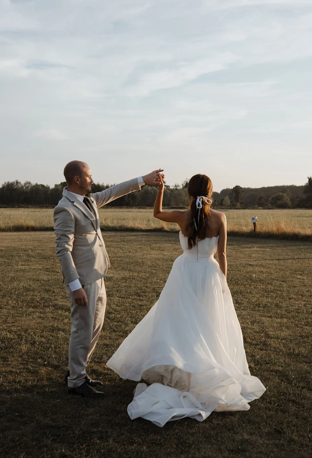 Bride and groom dancing outdoors in a field at sunset, with the bride in a white wedding gown and the groom in a light-colored suit.
