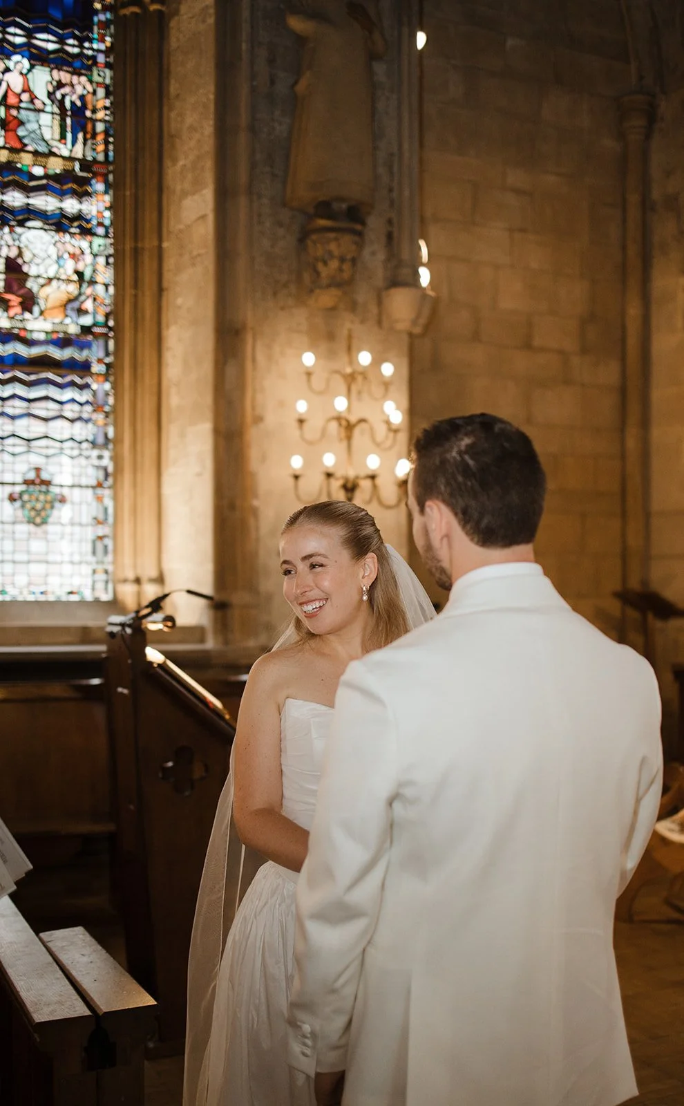 A bride and groom in a church during their wedding ceremony, standing near a stained glass window and a candelabra chandelier.
