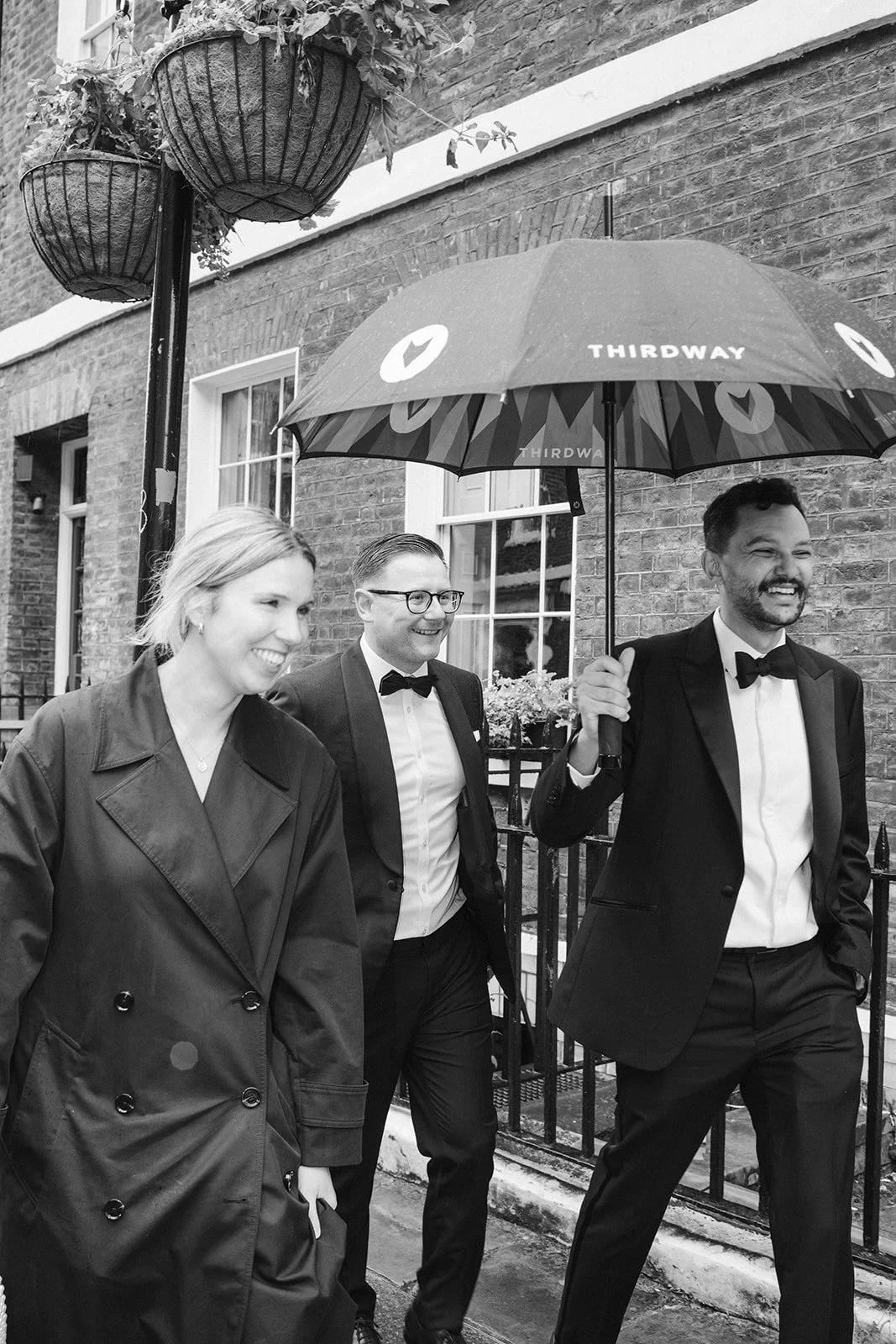 Three people in formal attire walking outside in the rain under an umbrella, with a brick building in the background. The man on the right is holding the umbrella, and all are smiling.