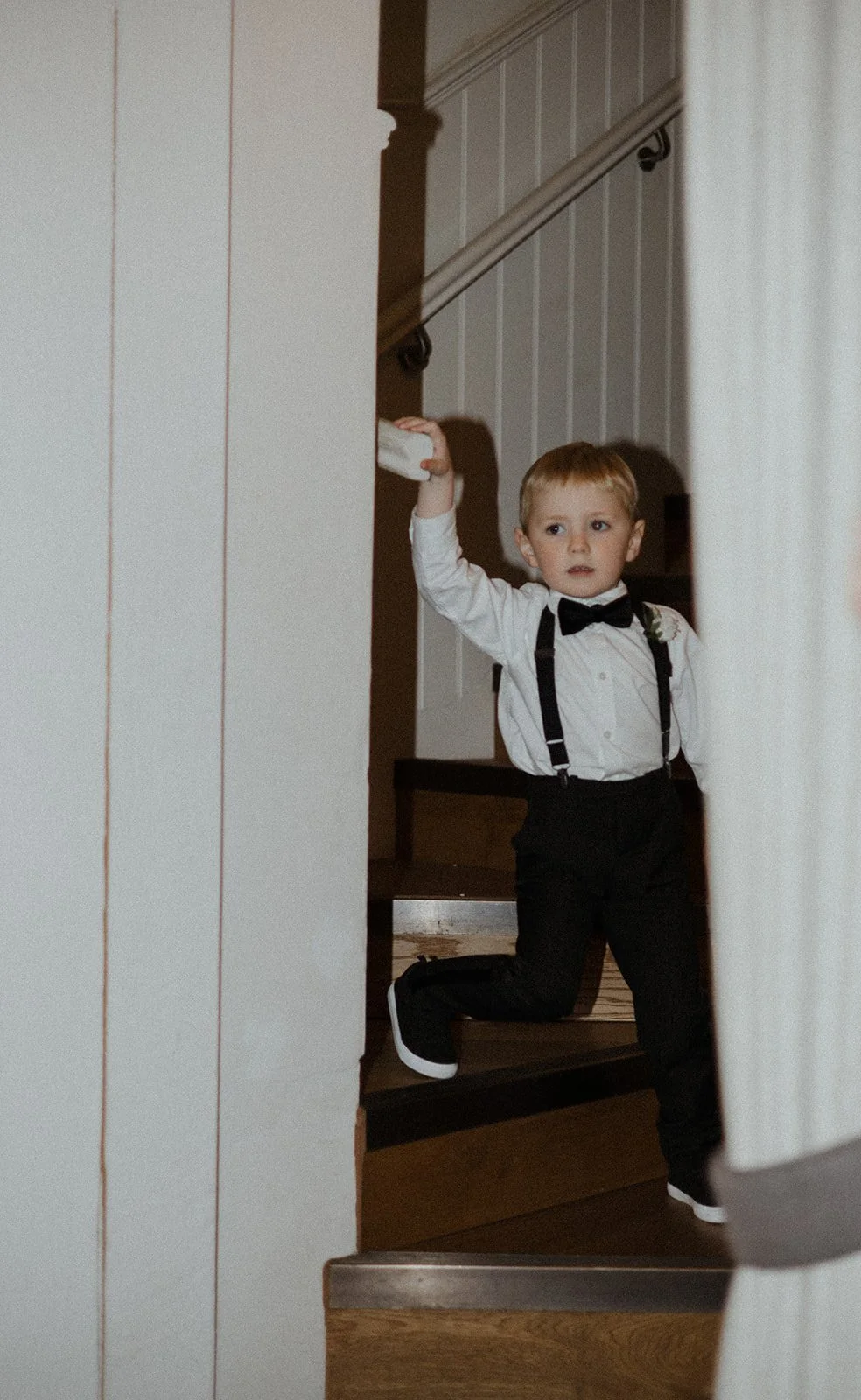 Young boy dressed in formal attire with a bow tie, suspenders, and black shoes, holding onto a staircase railing while kneeling on the stairs.