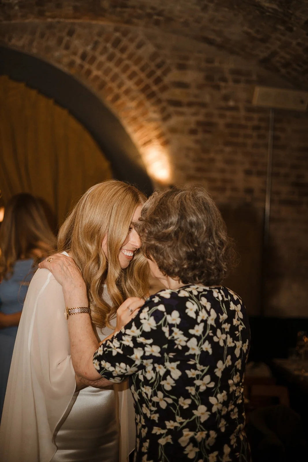 Two women sharing an intimate moment, smiling and leaning close, in a dimly lit brick-walled venue.