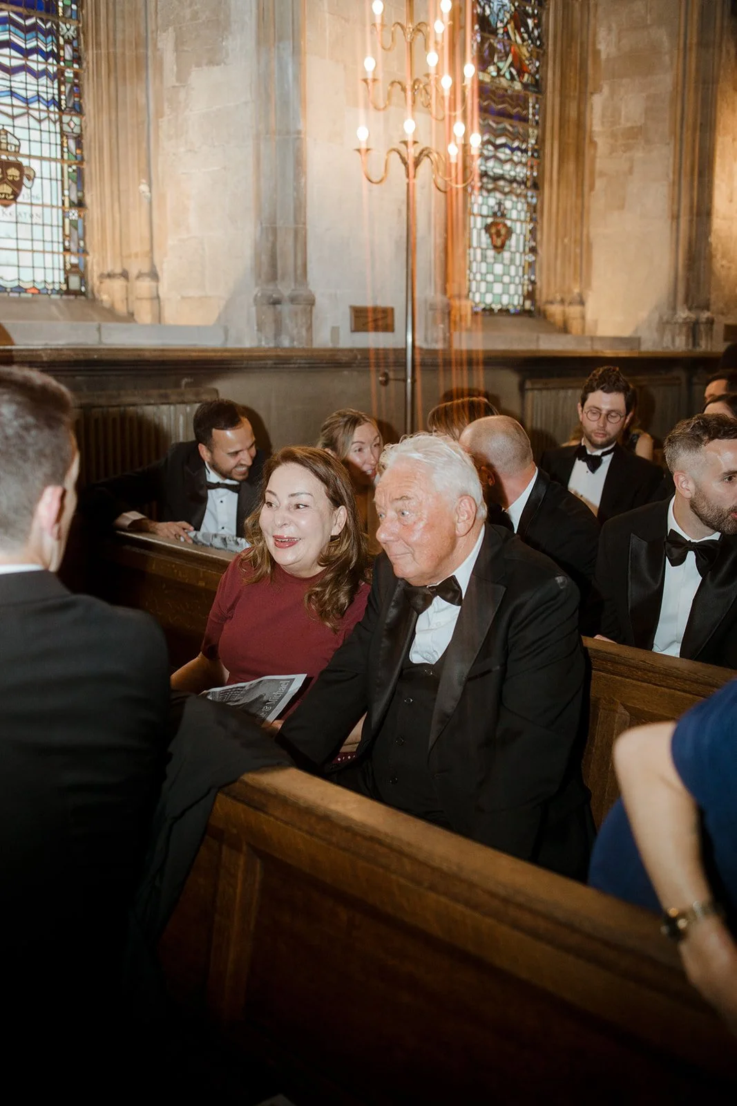 People dressed in formal attire sitting in church pews during a wedding or formal event, with stained glass windows and a chandelier in the background.