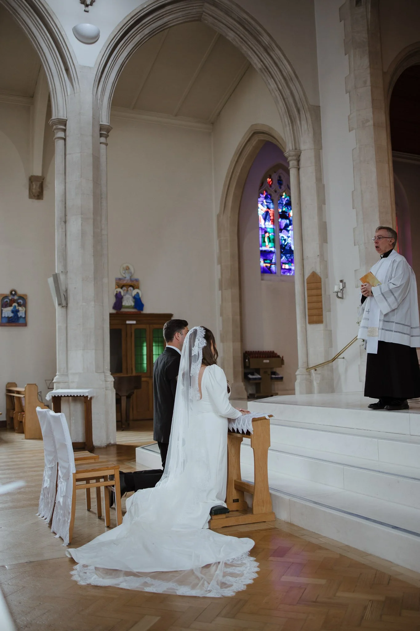 A bride and groom kneeling at the altar during a wedding ceremony inside a church, facing a priest standing on the altar, with stained glass windows behind him.
