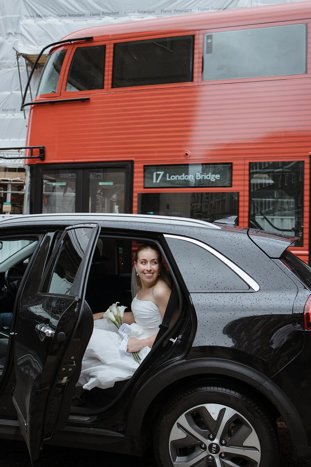 A woman in a white wedding dress sitting in the back seat of a black car, holding a bouquet of white calla lilies, smiling, with a red double-decker bus in the background displaying Destination 17 London Bridge.