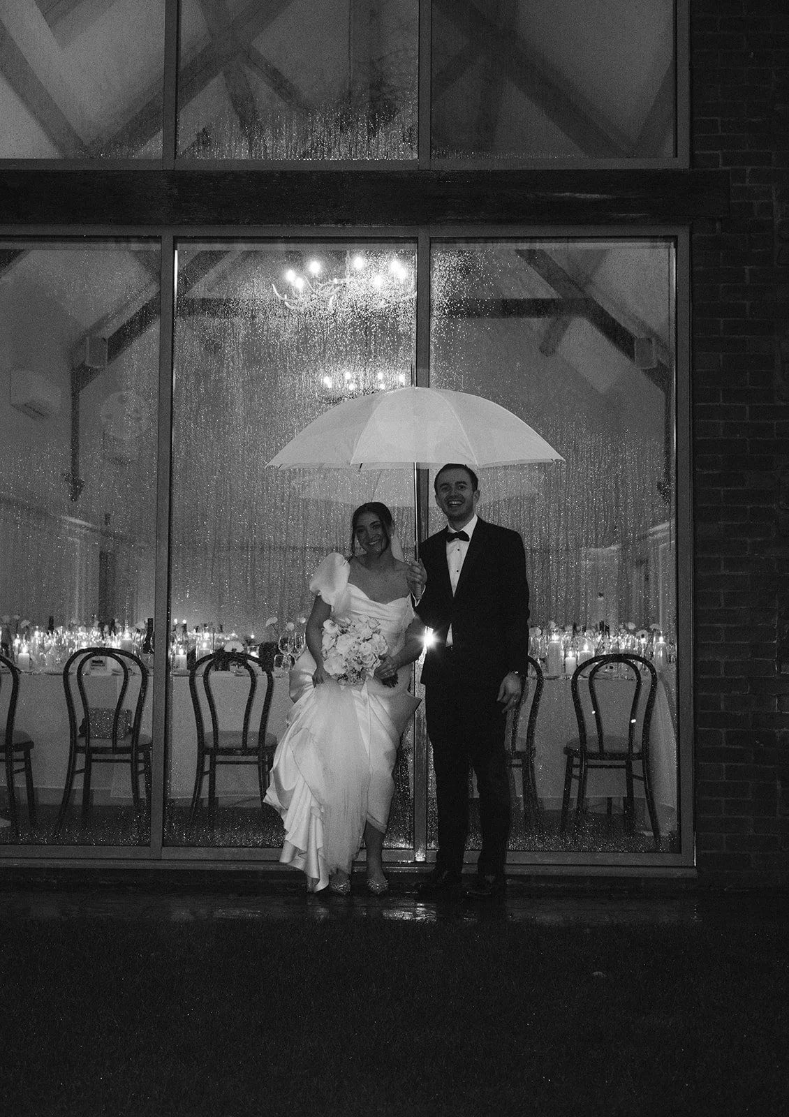 A wedding couple stands outside in the rain under an umbrella, with the bride holding a bouquet and the groom dressed in a tuxedo, in front of a large window showing a decorated interior.