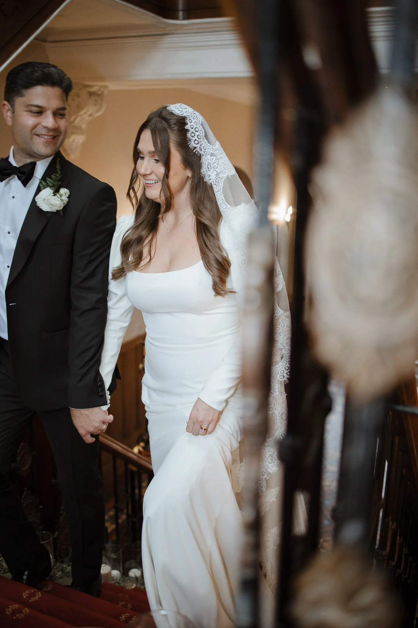 Bride and groom descending a staircase at their wedding, holding hands, with the bride wearing a white dress and lace veil, and the groom in a black tuxedo with a white boutonniere, smiling