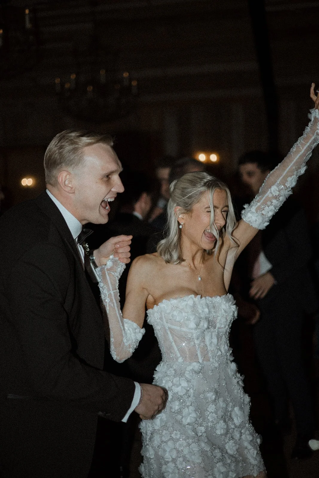 A bride and groom are dancing and celebrating at their wedding reception. The bride is wearing a white wedding dress with lace and floral details, and she has her arm raised with a joyful expression. The groom is dressed in a black tuxedo and is hold