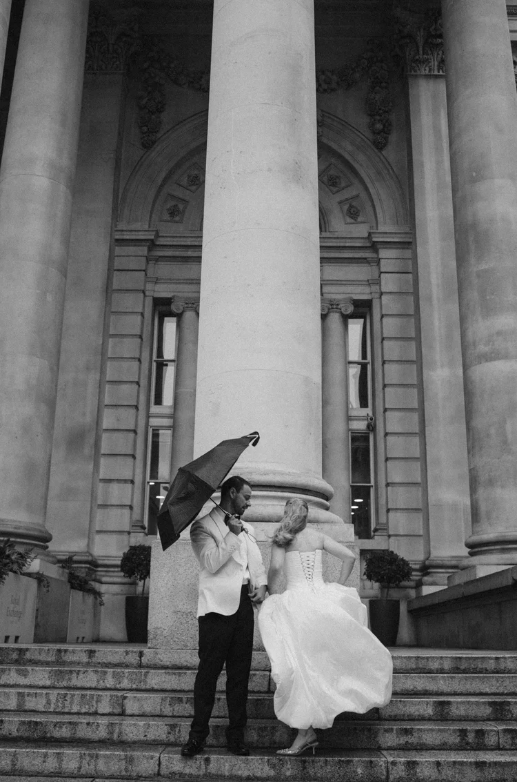 A man and woman in wedding attire standing on steps in front of a grand, classical building with large columns. The man holds an umbrella while the woman is adjusting her dress.
