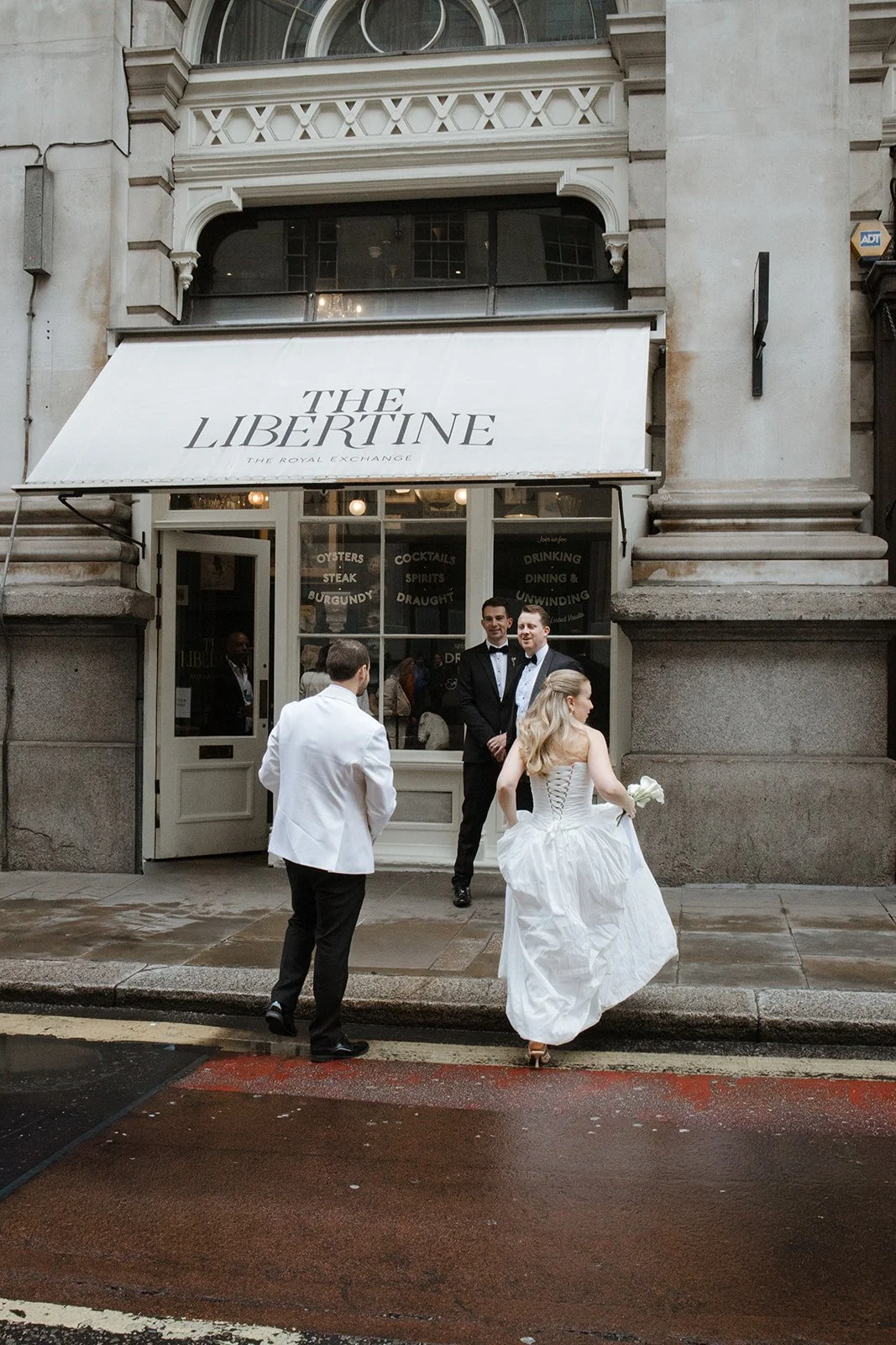 People dressed for a wedding outside The Libertine restaurant with a white awning and large glass windows in an urban setting.