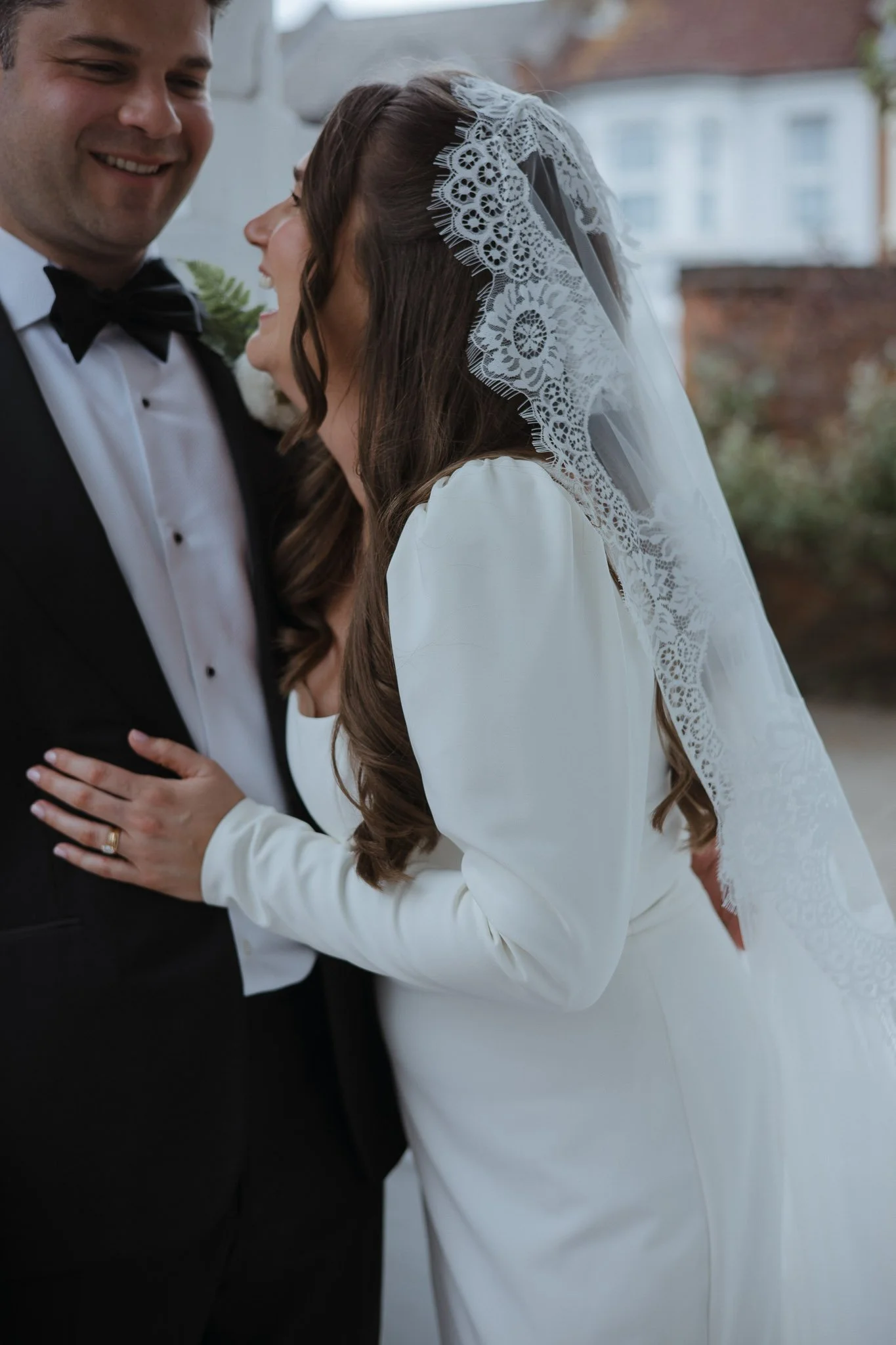A groom and bride smiling closely outdoors, the bride wearing a white dress and lace veil, the groom in a black tuxedo with bow tie.