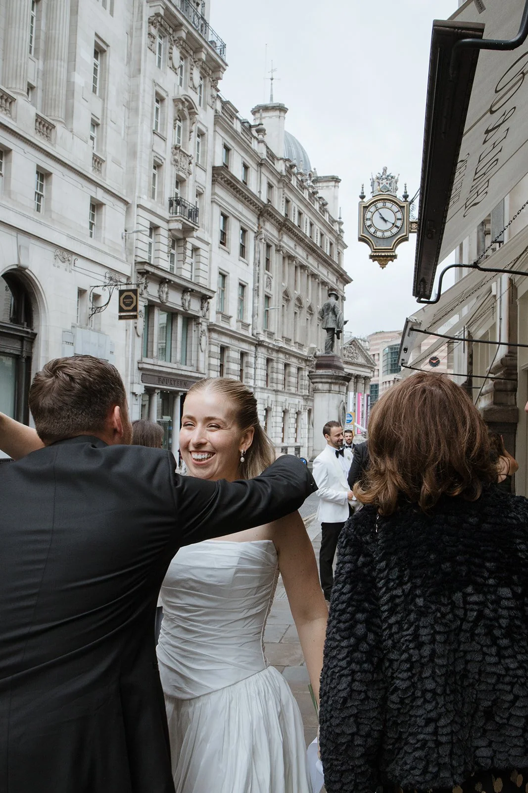 People celebrating outdoors on a city street, including a woman in a white dress smiling, with historic buildings and a clock in the background.