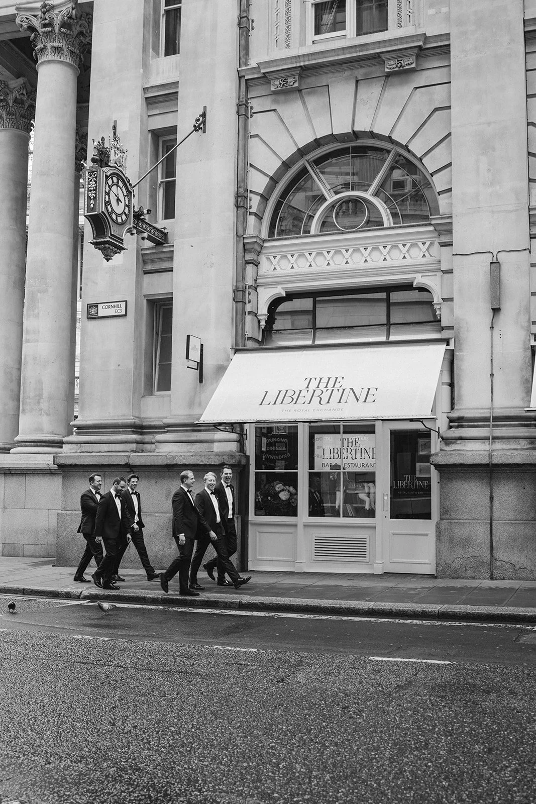 A group of seven men in tuxedos walking past the entrance of The Libertine restaurant, located on a city street with historic architecture, including columns and decorative window frames.