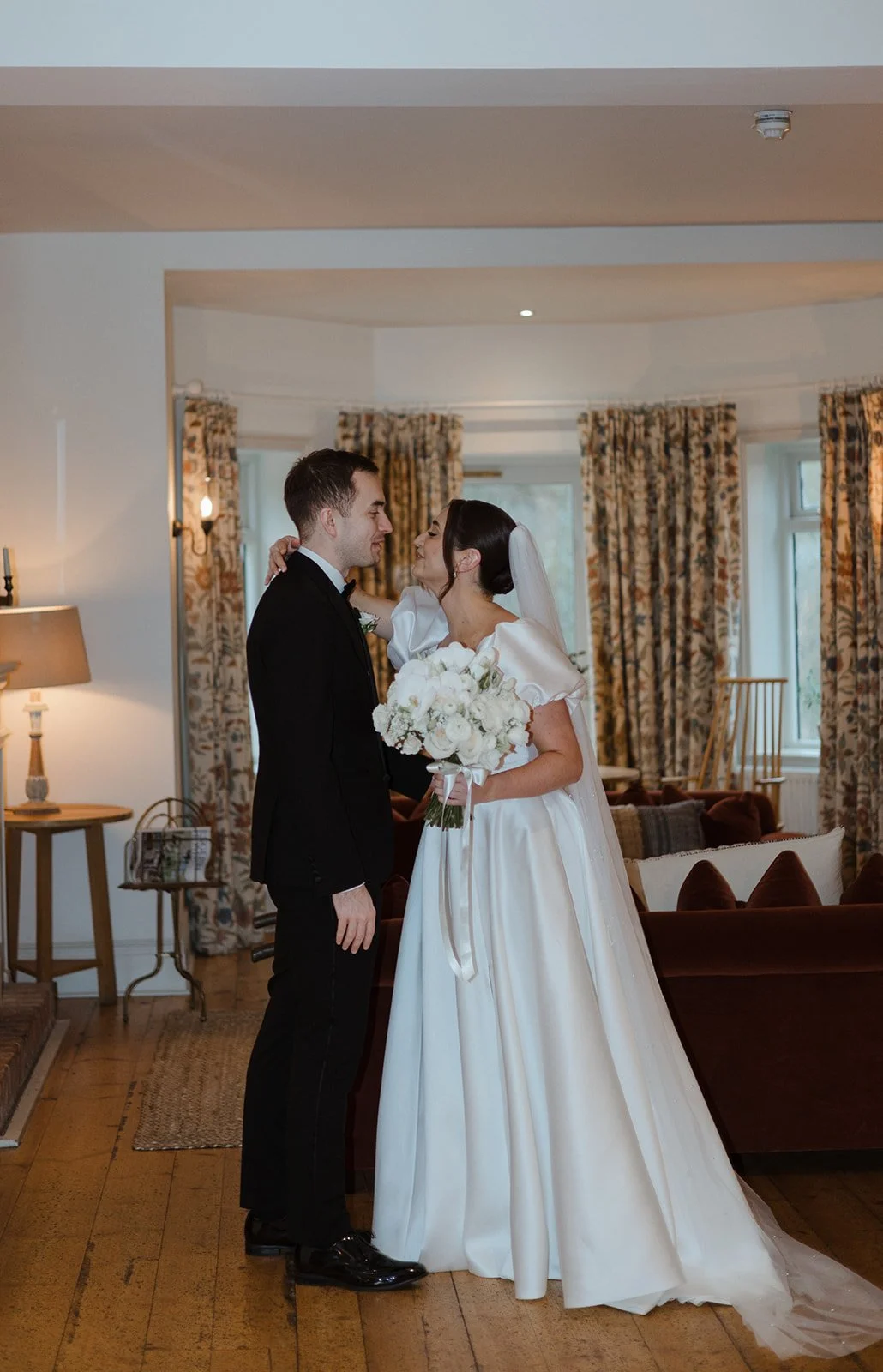 A bride and groom stand close together in a warmly lit living room, gazing into each other's eyes. The bride wears a white wedding gown and veil, holding a bouquet of white flowers. The groom is dressed in a black tuxedo. The background features flor