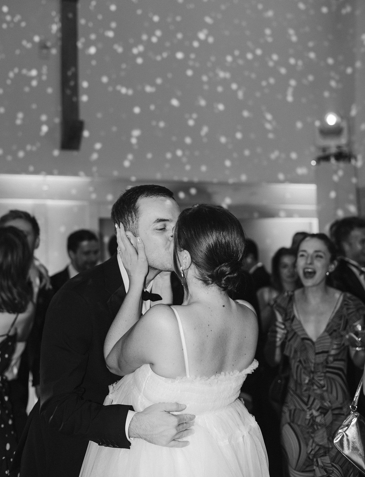 Black and white photo of a wedding reception with a bride and groom kissing, surrounded by onlookers, under decorated ceiling with hanging lights.