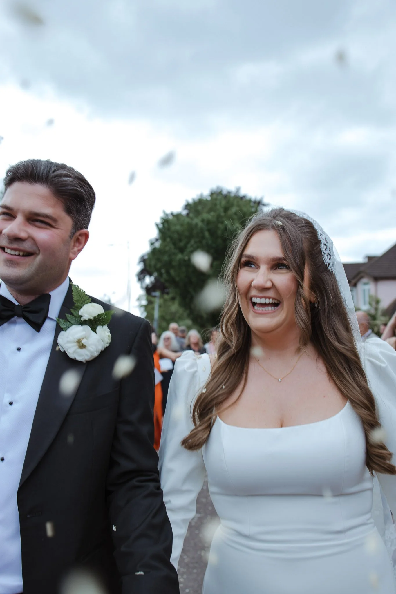 A smiling bride and groom walking outdoors during a wedding celebration, surrounded by guests and blowing confetti.