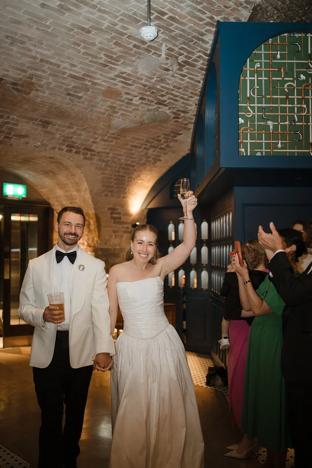 A newly married couple celebrating at their wedding reception, holding hands and raising glasses, with friends clapping and taking photos nearby, in an elegant venue with brick walls and arched ceilings.