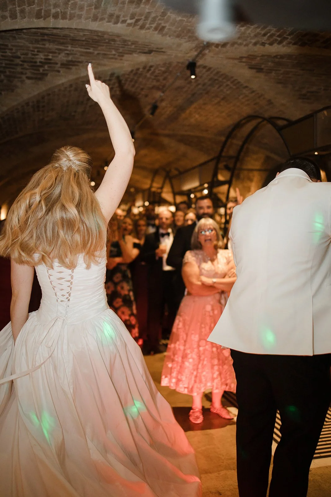 A bride in a white wedding dress with her arm raised at a wedding reception, with guests in formal attire in a brick-walled venue.