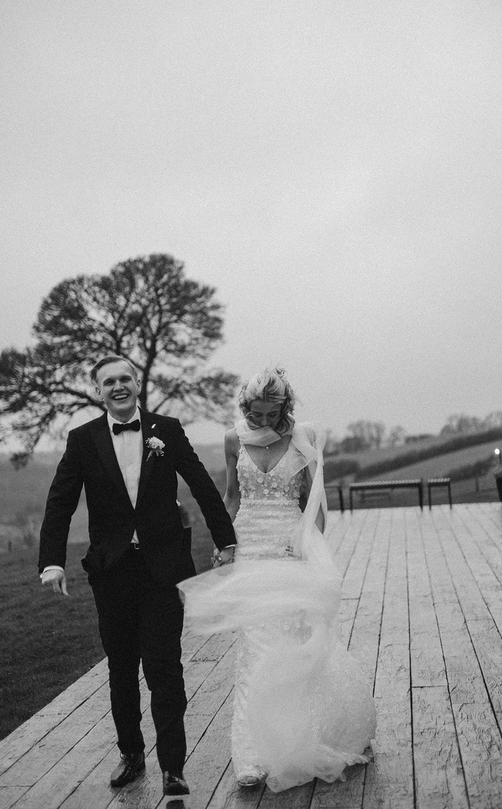 A black-and-white photo of a smiling groom and bride walking hand in hand on a wooden path outdoors, with trees and rolling hills in the background.