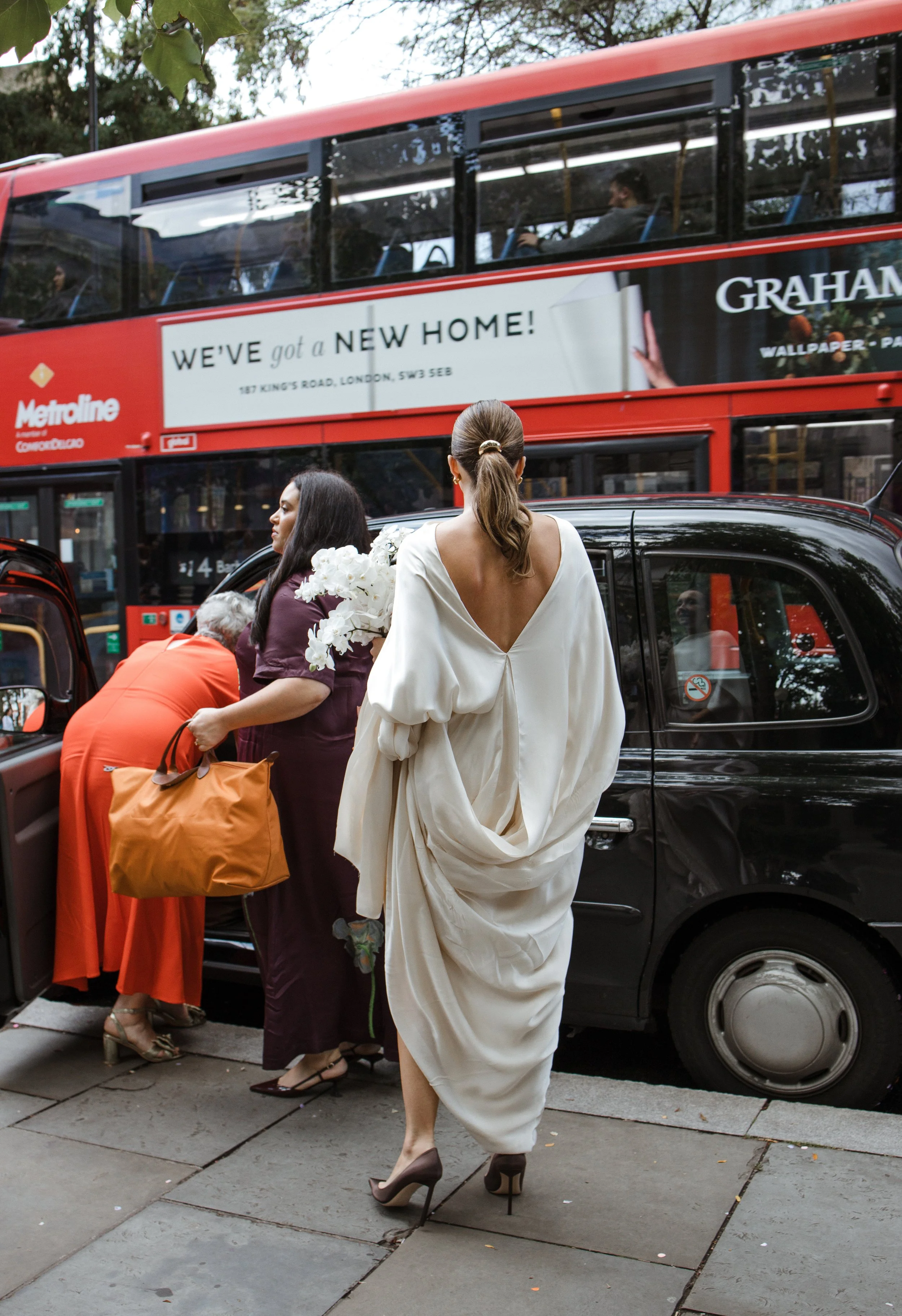 A woman in a white dress and high heels stands on the sidewalk with her back to the camera. Around her, people are getting into a black taxi. In the background, a red double-decker bus with an advertisement for a new home is passing by.