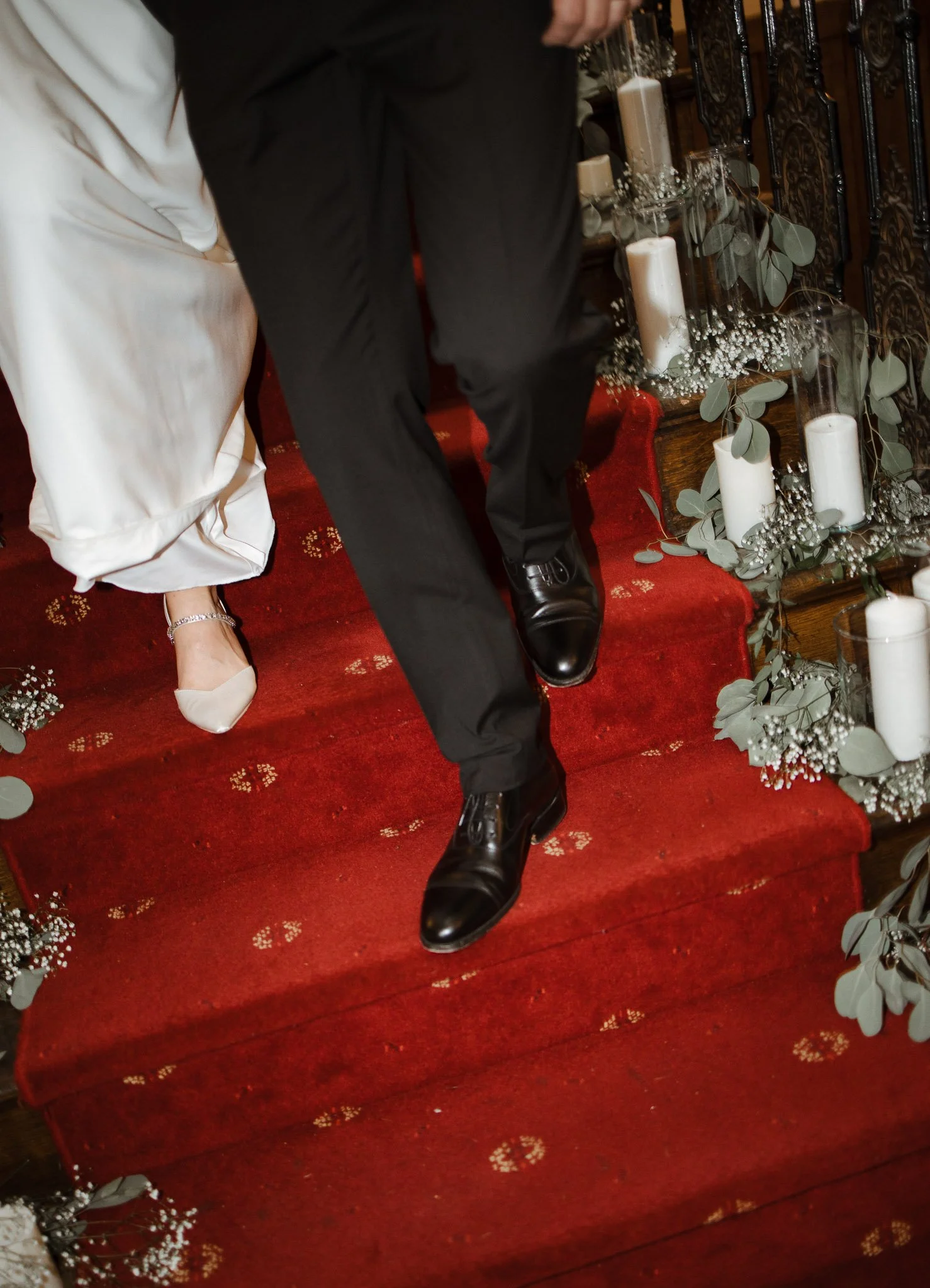 Close-up of a bride and groom holding hands as they walk down a red-carpeted staircase decorated with white candles, eucalyptus leaves, and baby's breath flowers during a wedding ceremony.