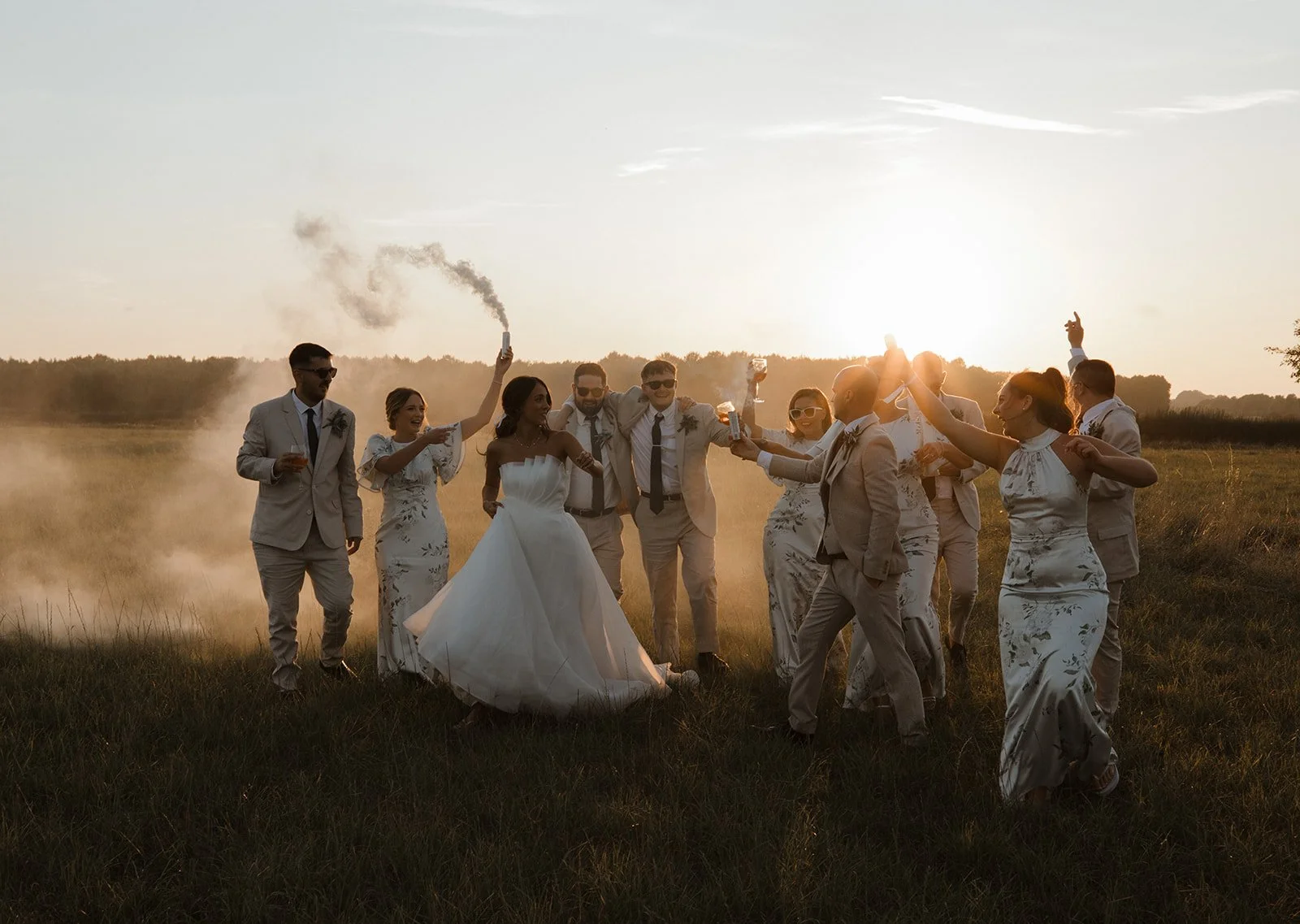 Wedding party celebrating outdoors at sunset, with the bride and groom in the center, surrounded by guests holding drinks and cheering.