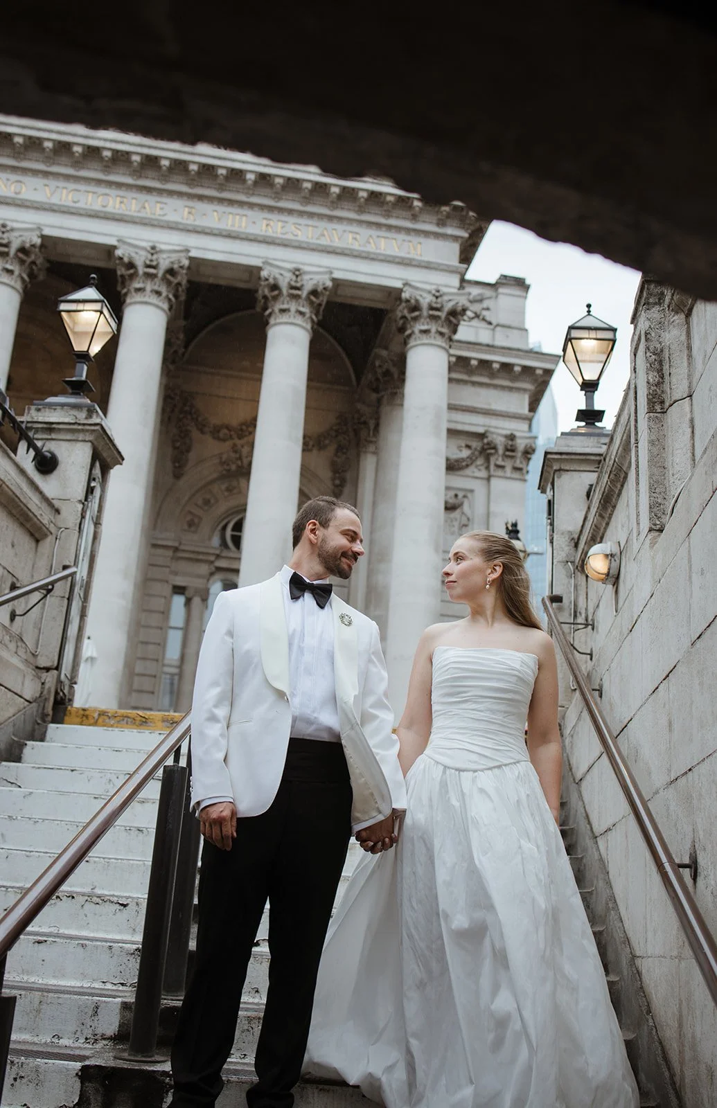 A couple in wedding attire holding hands on the stairs outside a historic building with classical columns and ornate details.