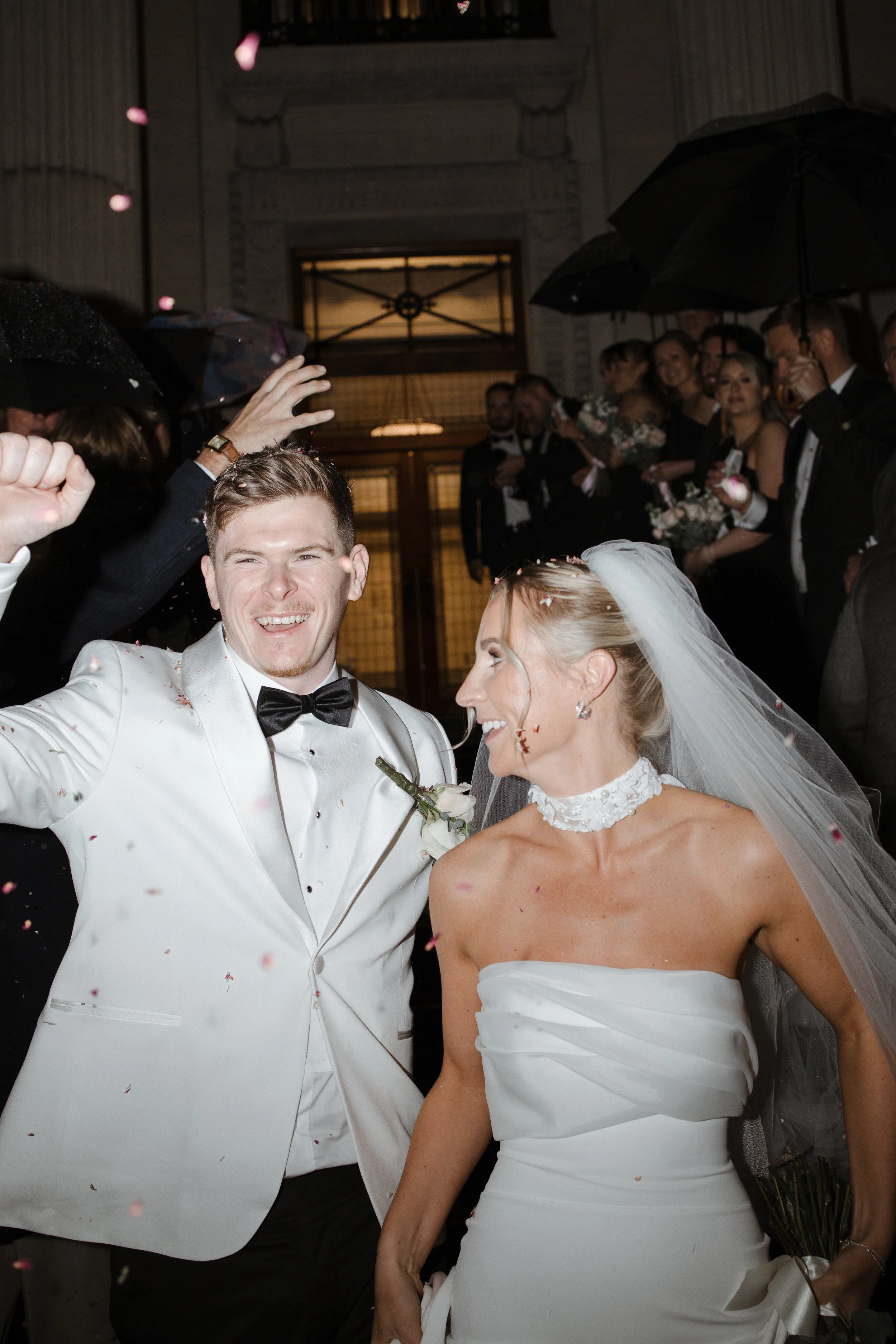 A newlywed couple on their wedding day, smiling and surrounded by guests holding umbrellas in a nighttime celebration, with pink flower petals falling.