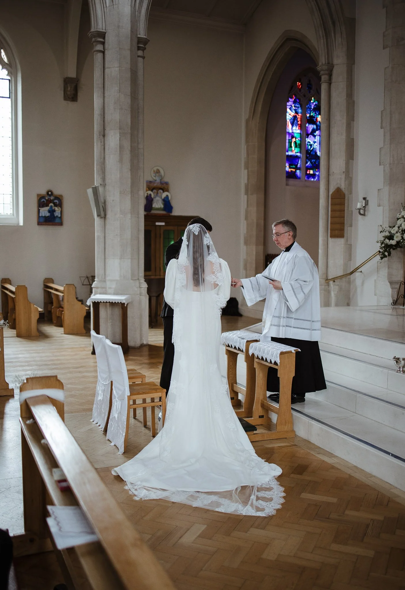 A bride and groom exchange wedding vows at the altar inside a church, with a priest officiating, stained glass windows, and wooden pews.