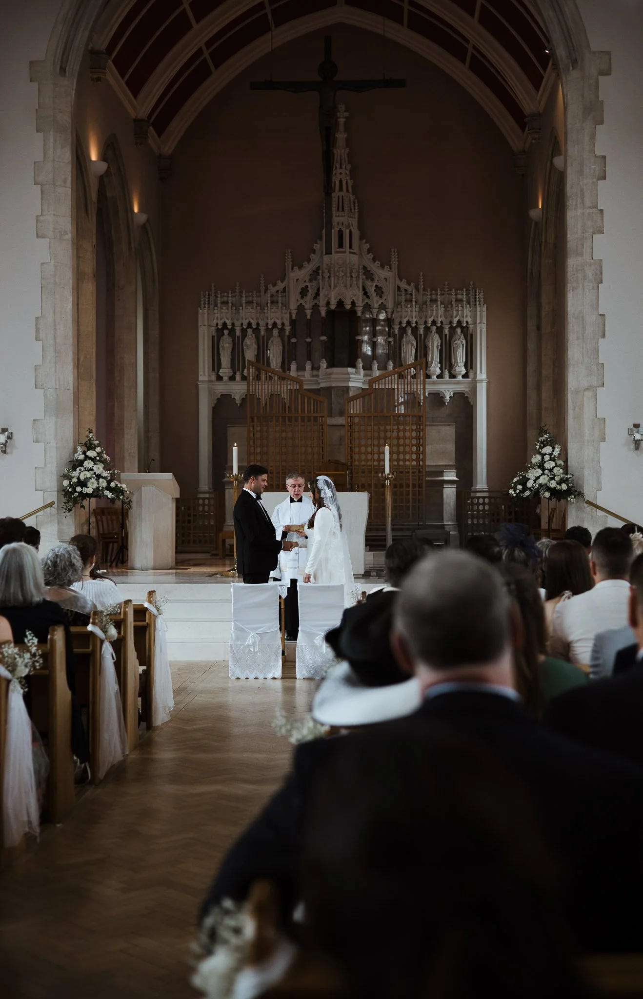 A couple gets married at the altar during a wedding ceremony inside a church, with a priest officiating and guests seated on benches.