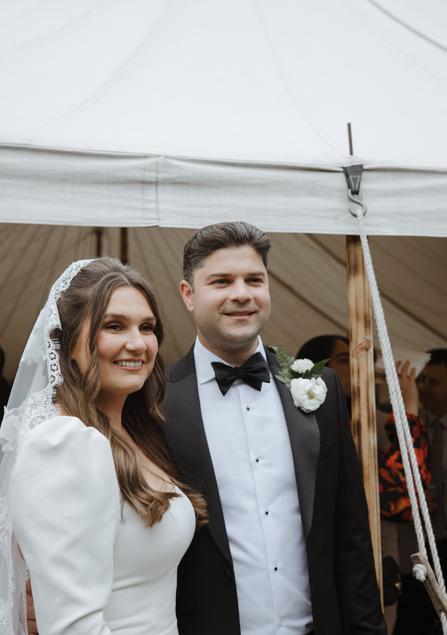 A smiling bride and groom dressed in wedding attire standing under a tent, with the bride wearing a white dress and lace veil and the groom in a black tuxedo with a bow tie and boutonnière, at their wedding ceremony.