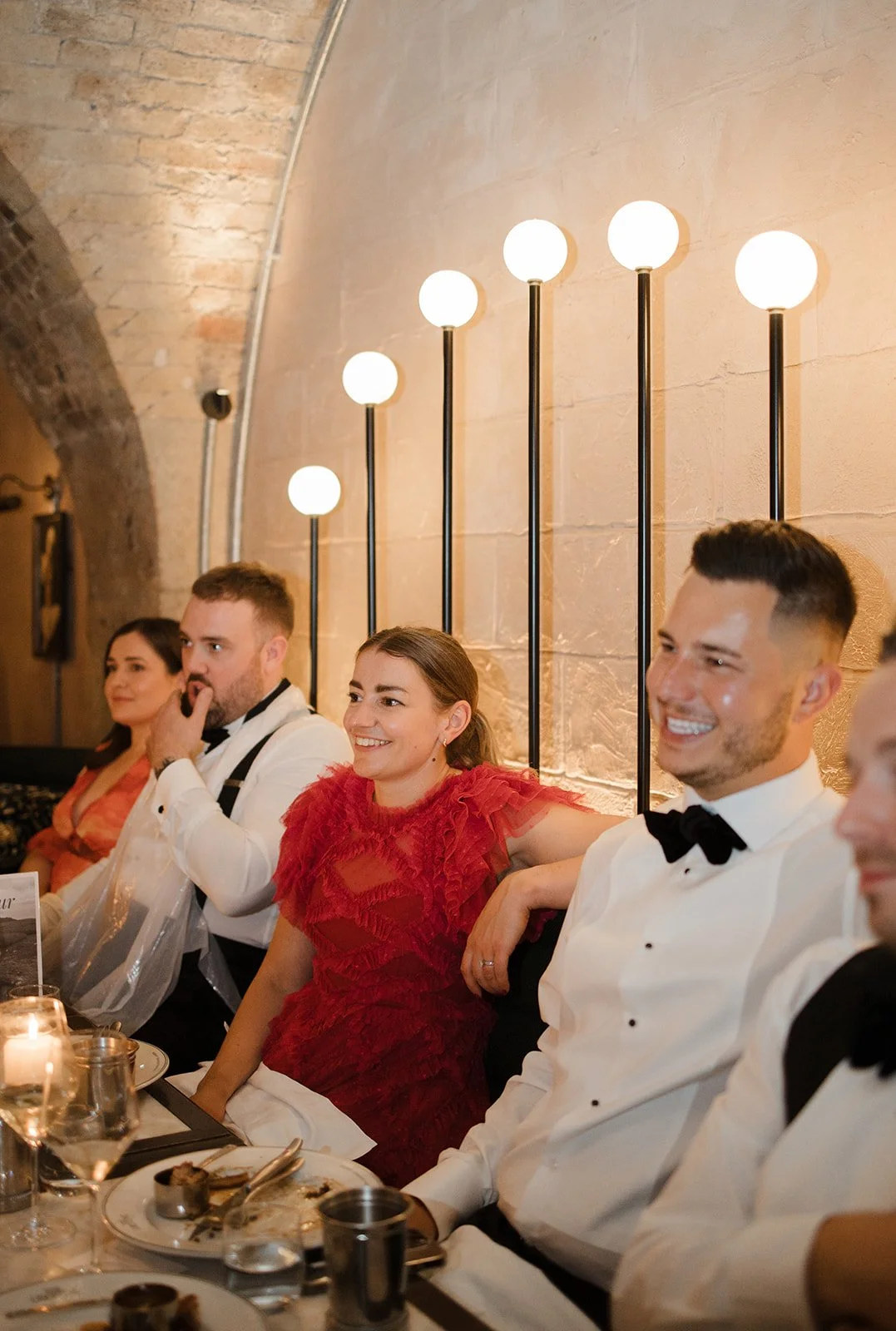People dressed in formal attire are sitting at a long dinner table in a warmly lit room with brick arch ceiling, smiling and enjoying a celebration.