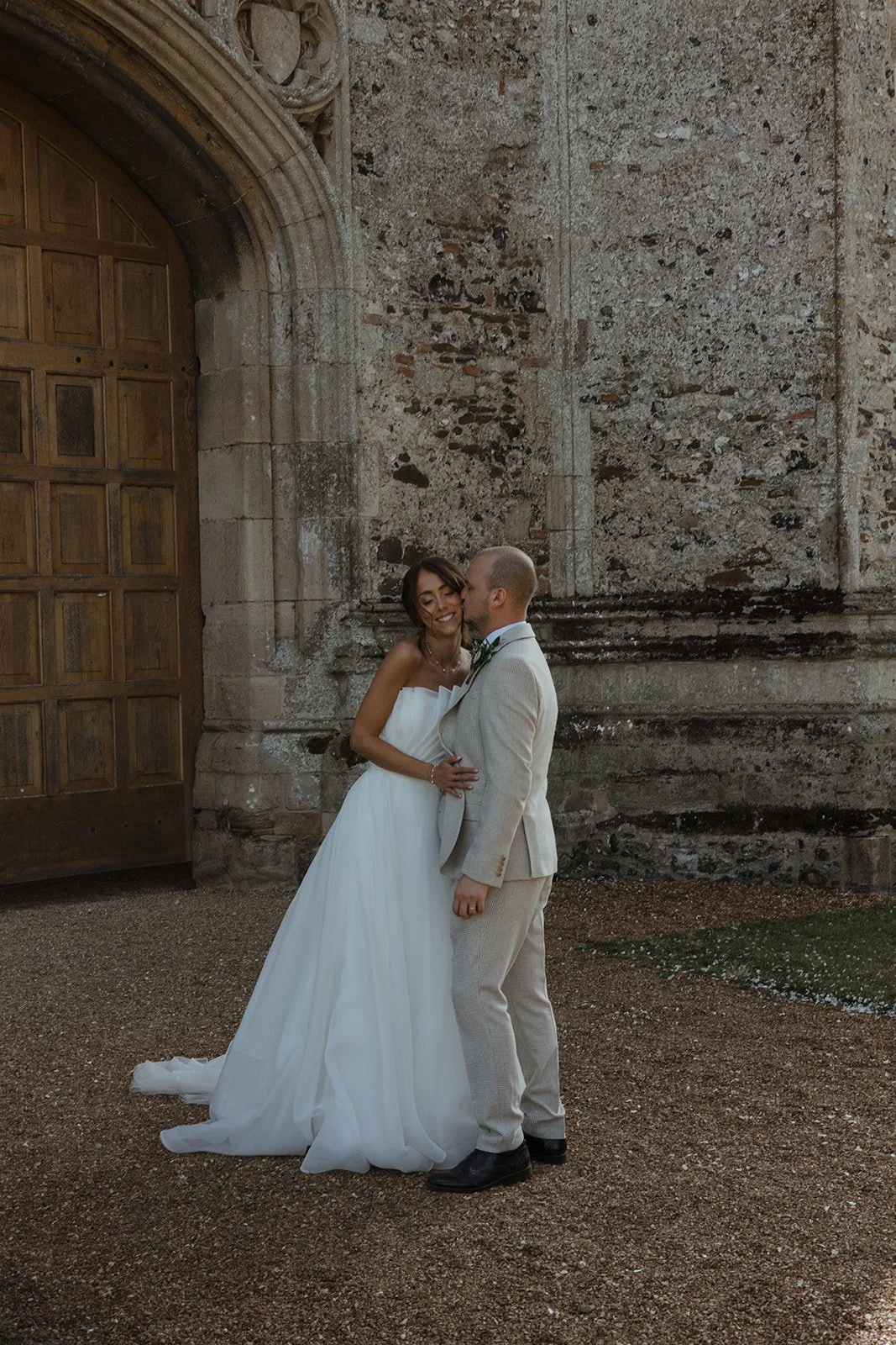 A bride and groom are embracing in front of an old stone building with a large wooden door, the bride wearing a white strapless wedding dress and the groom in a beige suit with a bow tie.