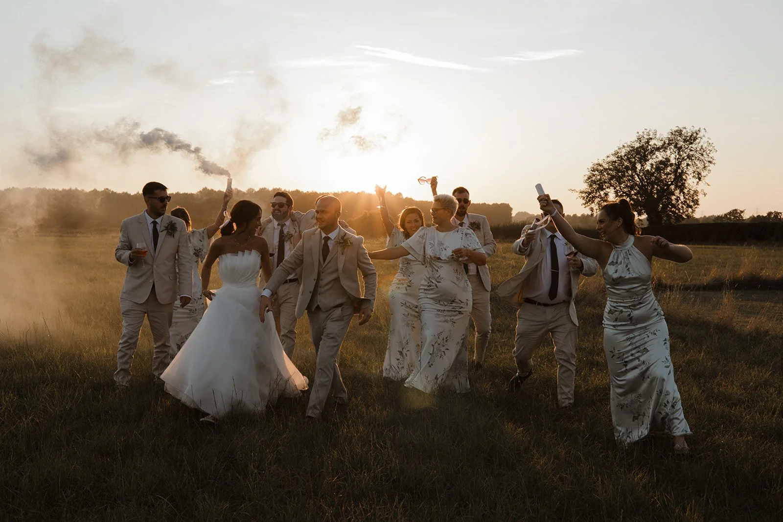A wedding party in formal attire celebrating outdoors at sunset, with some holding sparklers and a smoke flare, on a grassy field.