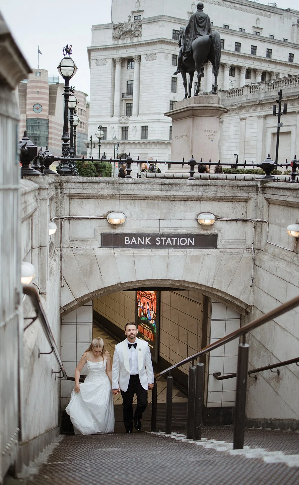A man and a woman in wedding attire walking up the stairs of a subway entrance labeled 'Bank Station' in London. The man is wearing a white tuxedo jacket with black pants and bow tie, while the woman is in a white wedding dress. There are buildings a