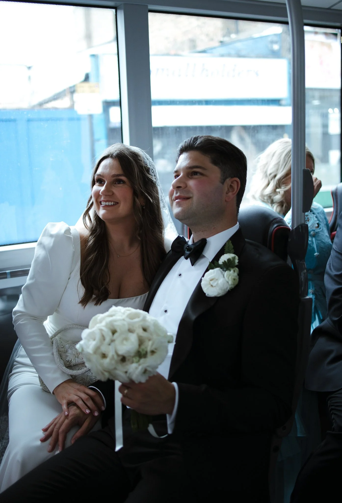 A bride and groom sitting together at their wedding, with the groom holding a bouquet of white flowers and the bride smiling, inside a venue with large windows.