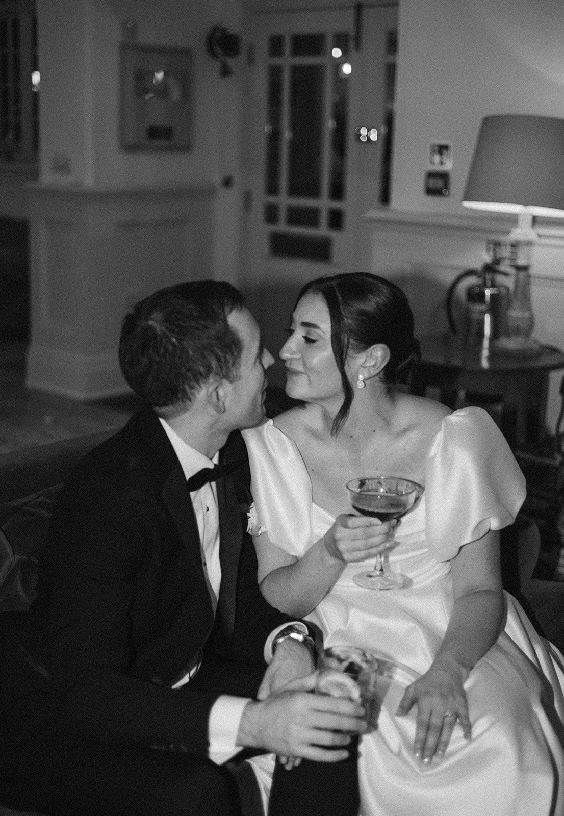 A black and white photo of a man and woman sitting close together, appearing to be about to kiss, with the woman holding a cocktail glass. They are dressed formally, the man in a tuxedo and the woman in a dress with puffed sleeves, in a cozy indoor s