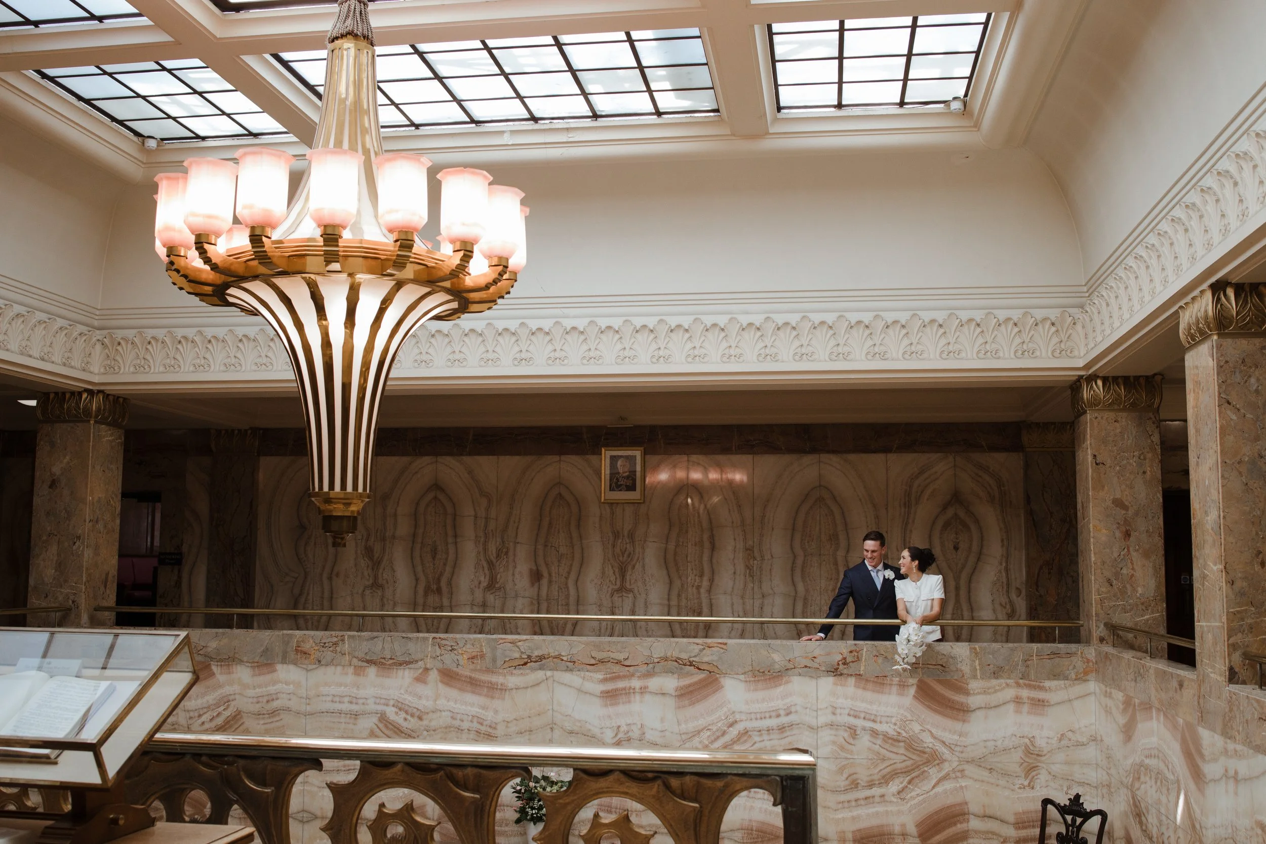 A chandelier hanging from a glass ceiling in an elegant lobby with marble columns and walls. Two people are standing in the background, smiling and talking.