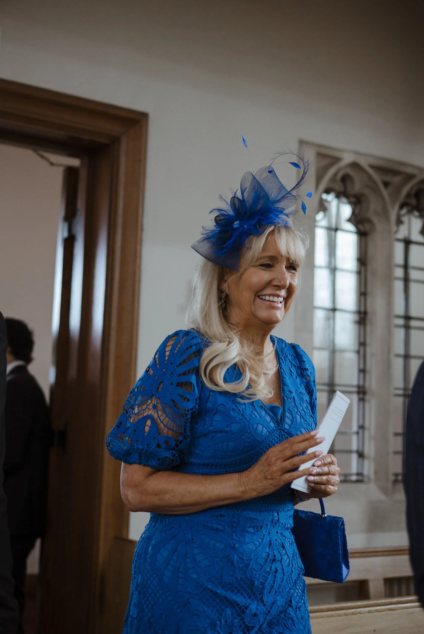 A smiling woman in a vibrant blue lace dress with matching fascinator, holding a program, inside a historic building with large stained glass windows.