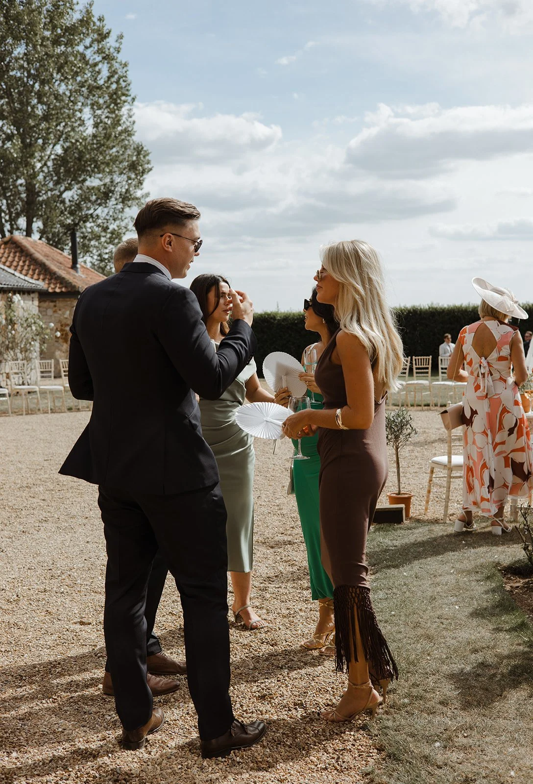 People socializing at an outdoor event on a sunny day, some holding wine glasses, with chairs and trees in the background.