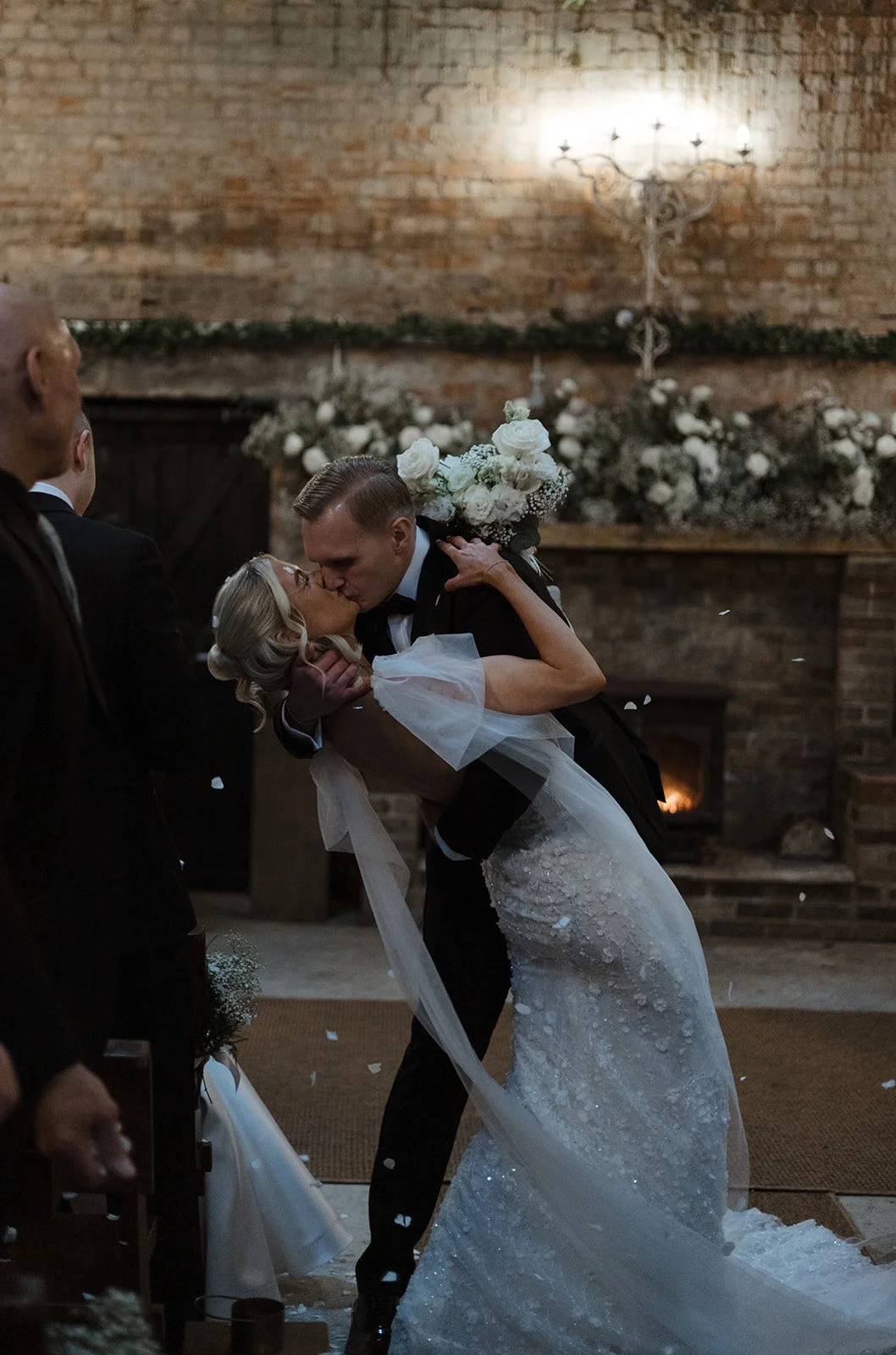 A bride and groom share a kiss during their wedding ceremony, with the groom dipping the bride. The bride is in a white wedding dress holding a bouquet of white flowers. The background features a brick wall with a candelabra and floral decorations.