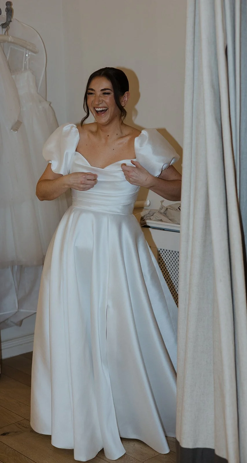 A woman wearing a white wedding dress, smiling and adjusting her dress, standing in front of a mirror.