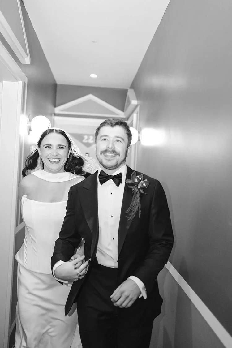 A black and white photo of a smiling bride and groom walking down a hallway, the bride wearing a white wedding dress and veil, and the groom in a black tuxedo with a bow tie and boutonniere.