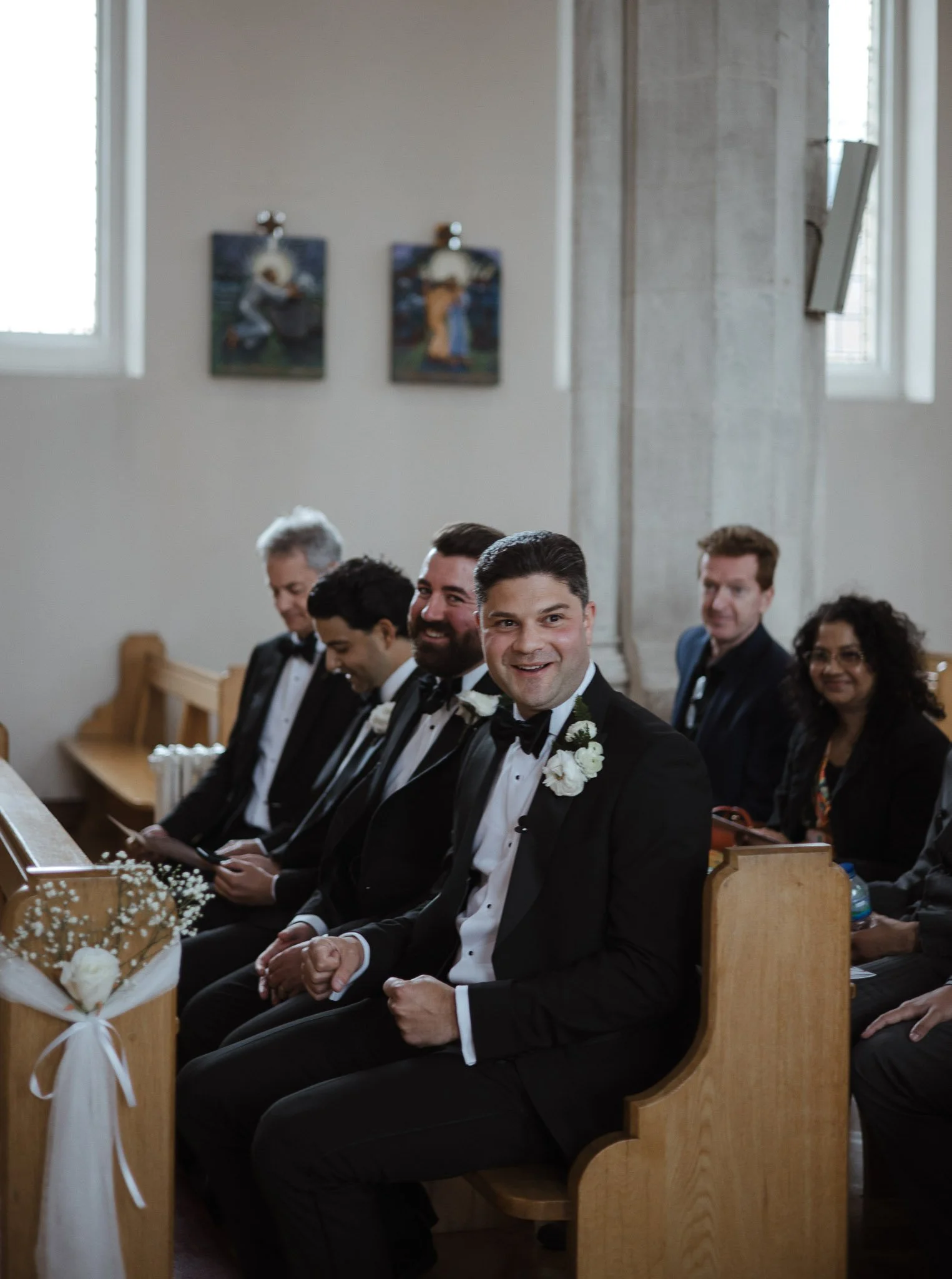 Group of men in tuxedos and women in formal attire sitting in pews inside a church, during a wedding ceremony.