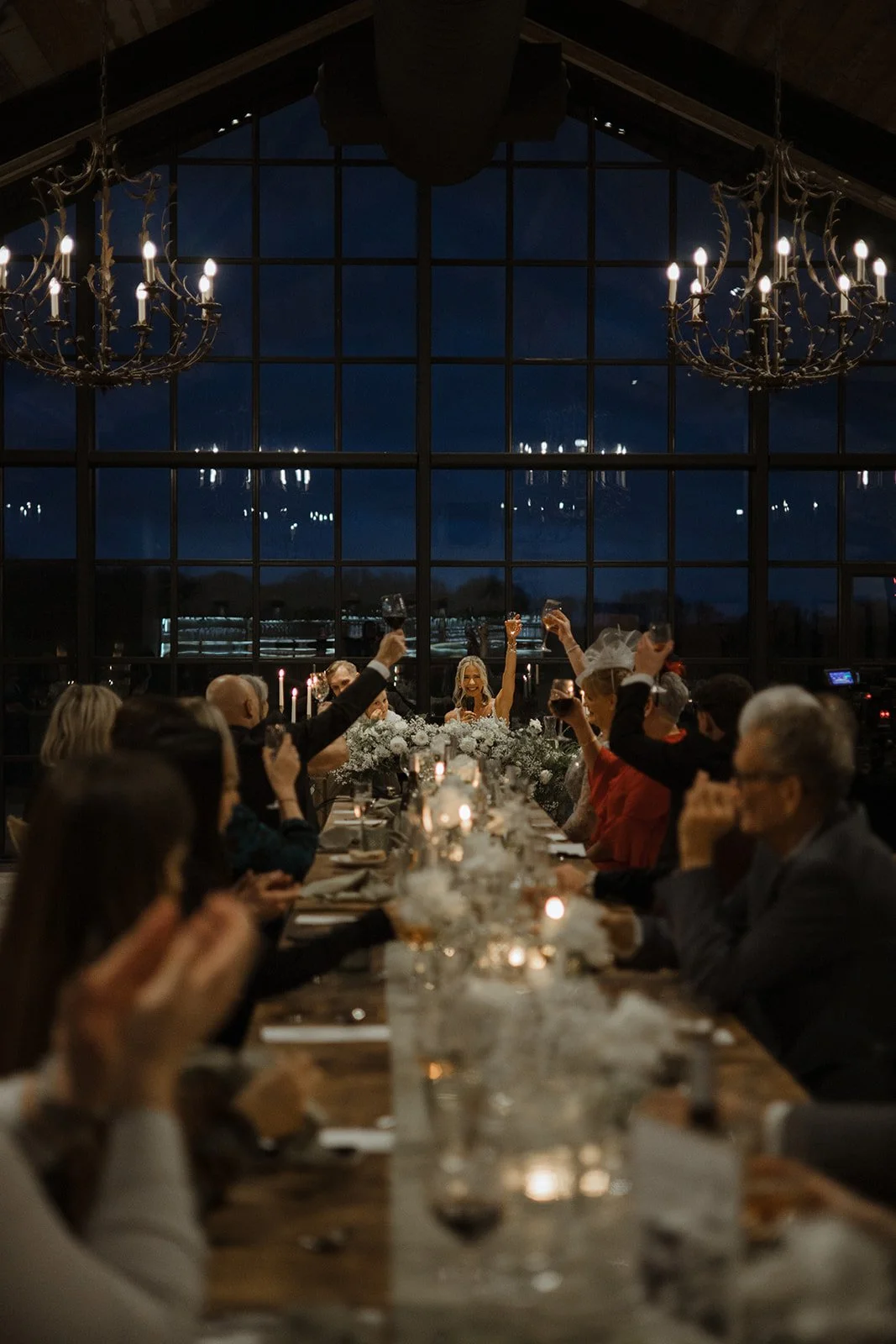 A nighttime dinner party with guests raising glasses in a toast at a long table decorated with white flowers and candles, inside a venue with large windows and chandeliers.