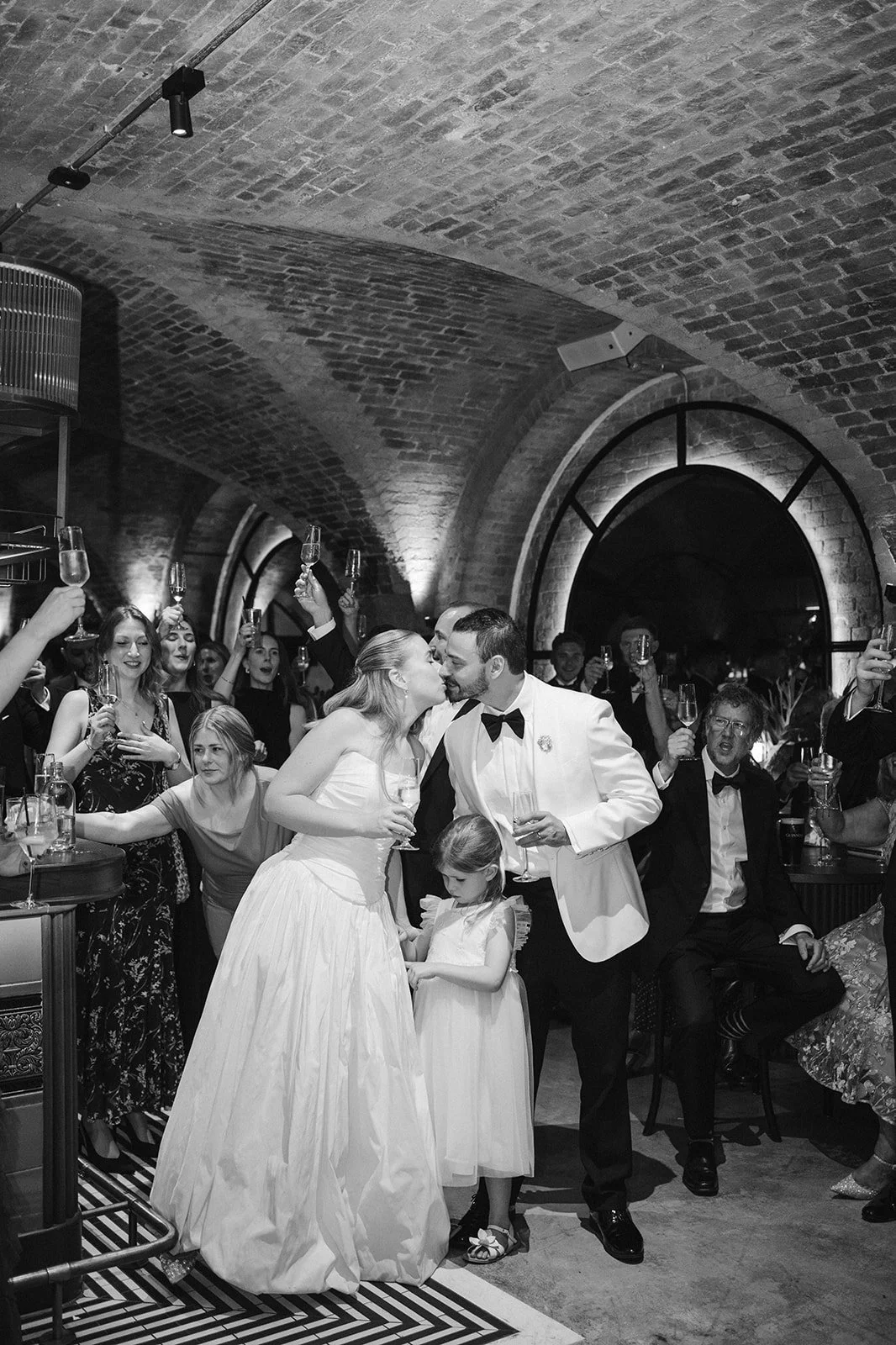 A wedding reception with the bride and groom sharing a kiss while guests celebrate around them, raising glasses in a celebration inside a brick archway.