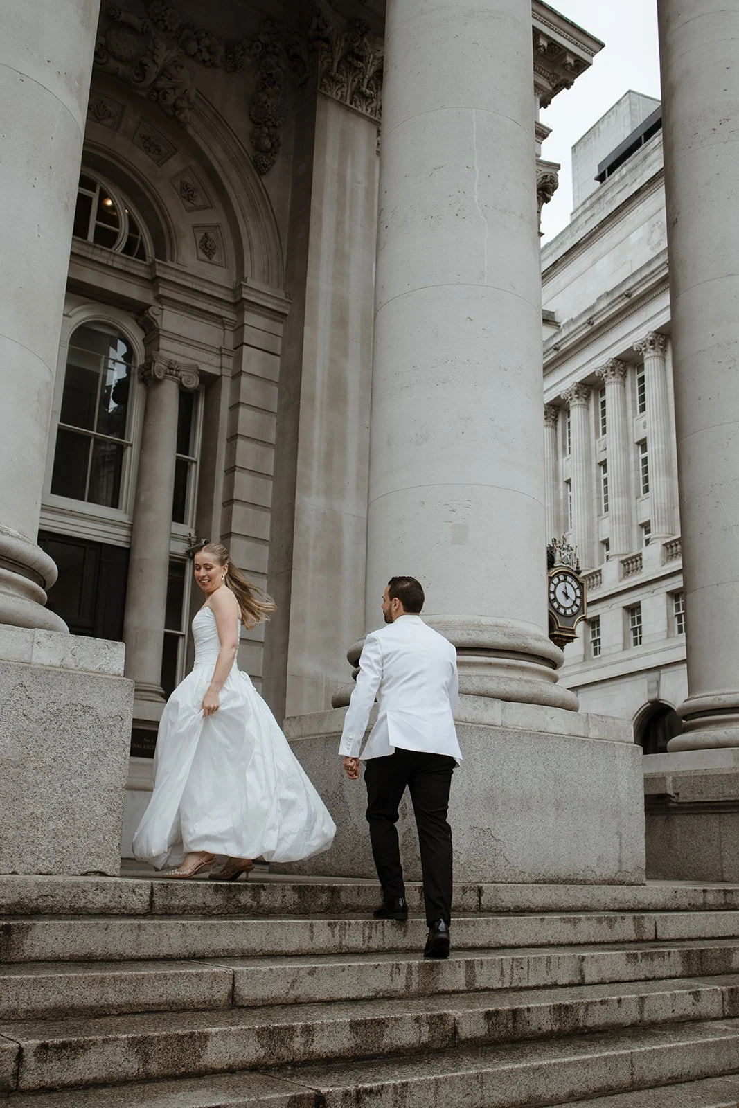 A bride in a white wedding gown and a groom in a white tuxedo jacket walking down the steps of a grand, historic building with large stone columns and a clock, possibly a courthouse or government building.