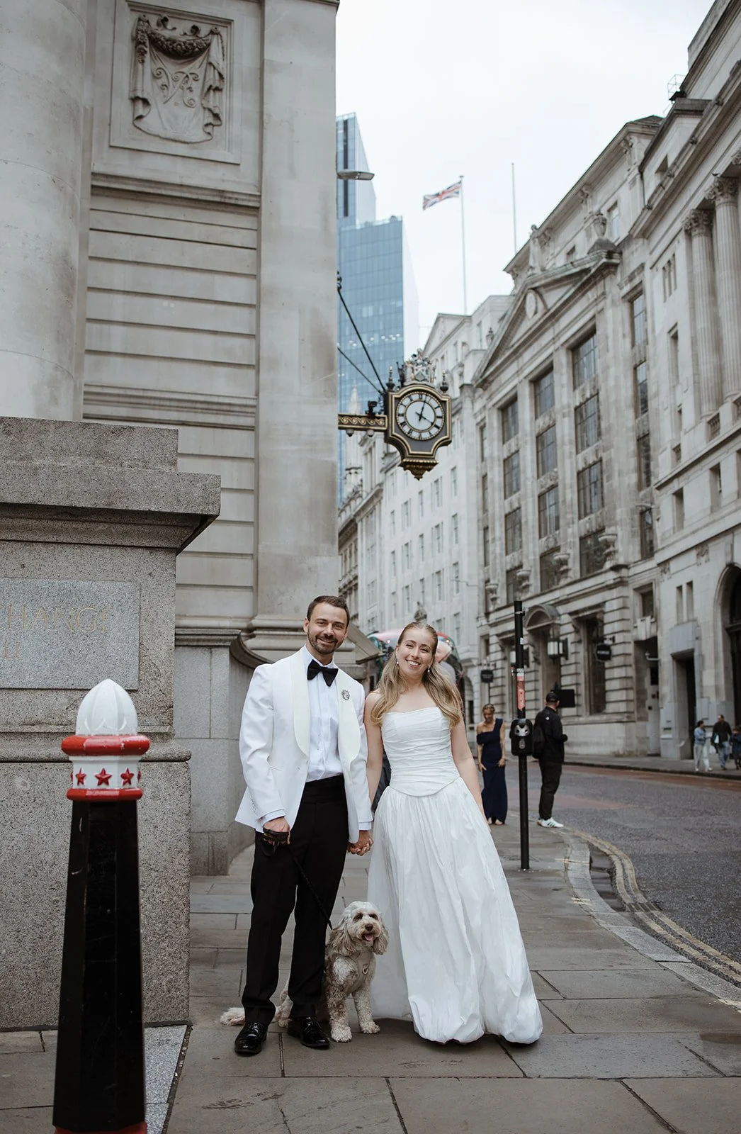 A newlywed couple in wedding attire, holding a dog, standing on a city street with historic buildings and a clock on a building behind them, during daytime.