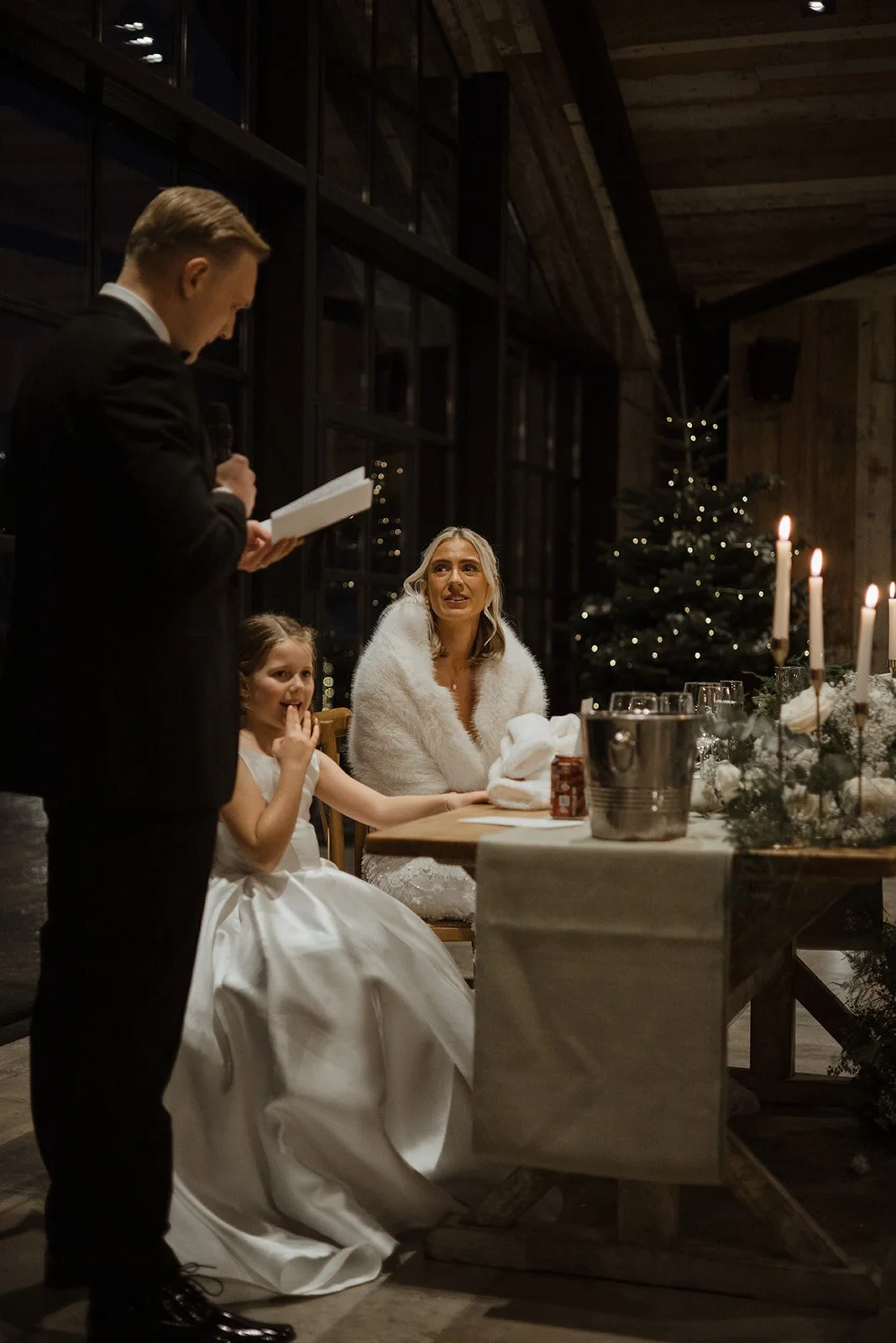A woman and young girl sit at a decorated dinner table during a Christmas celebration, while a man reads from a book nearby.