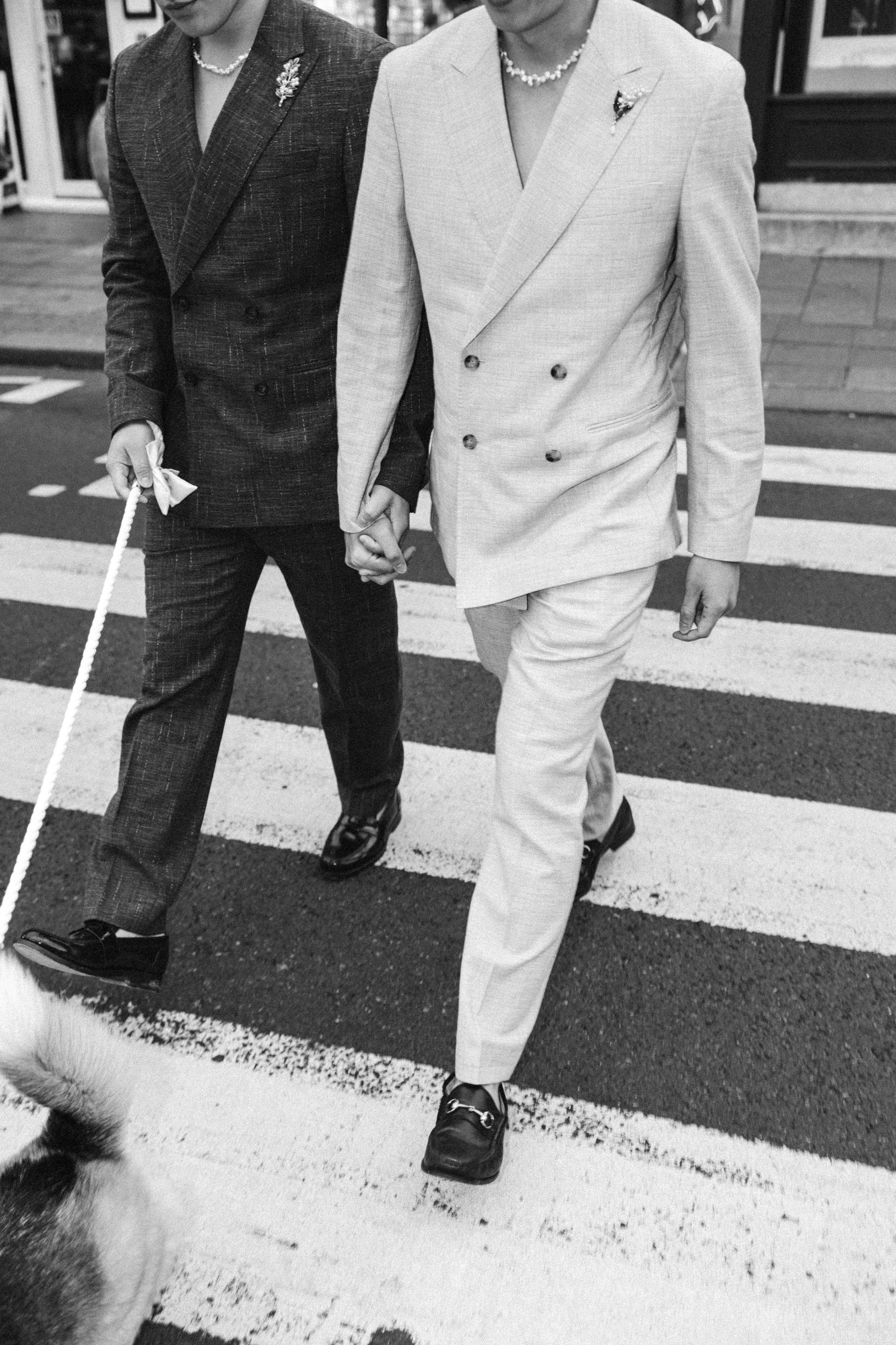 Two people walking hand in hand across a pedestrian crosswalk. One person is dressed in a light-colored suit, and the other in a dark suit. They are holding hands and wearing necklaces.